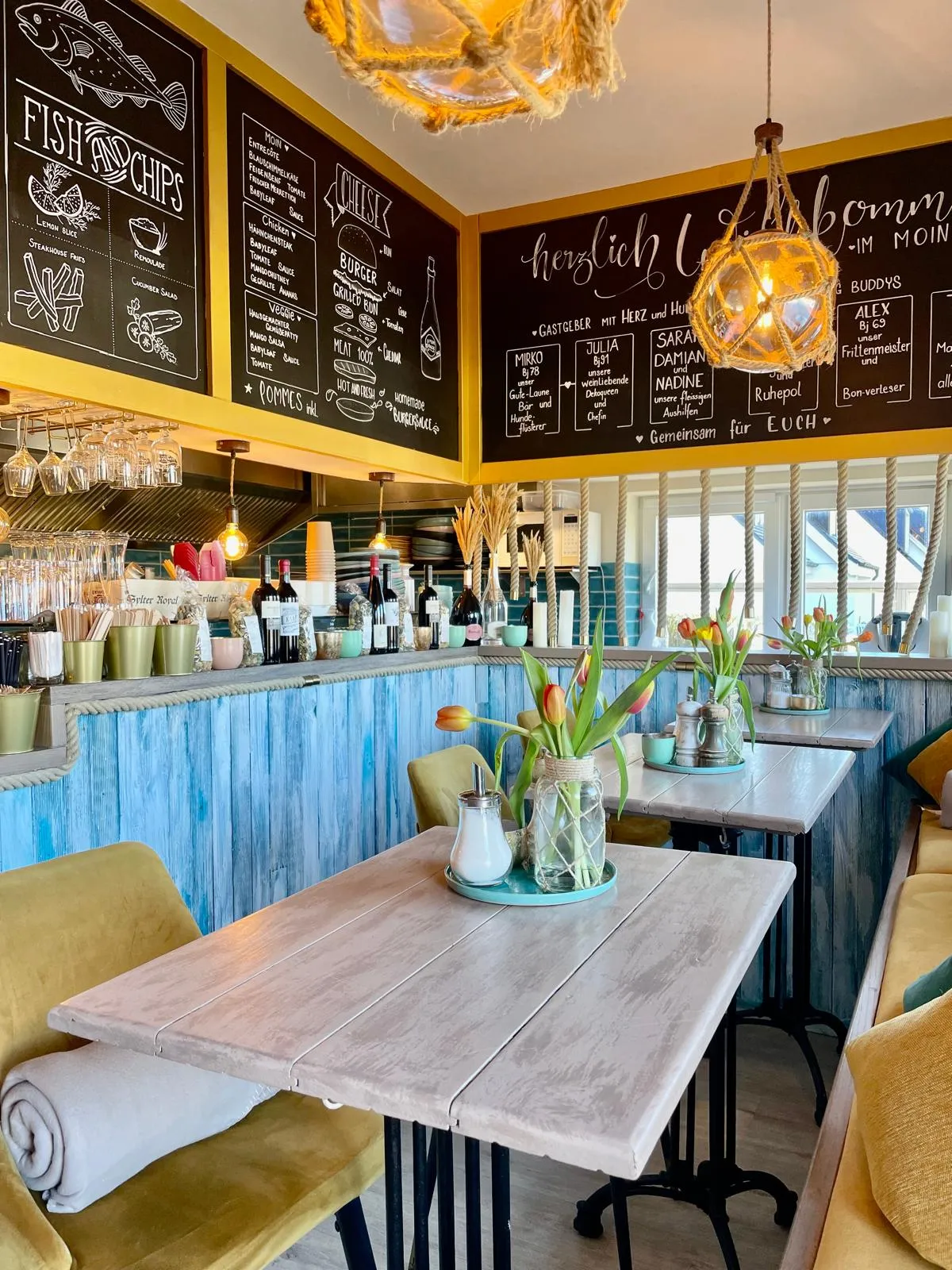 Helles Café mit maritimer Dekoration, blauen Wänden, Tischen mit Blumen und einer Getränketheke.