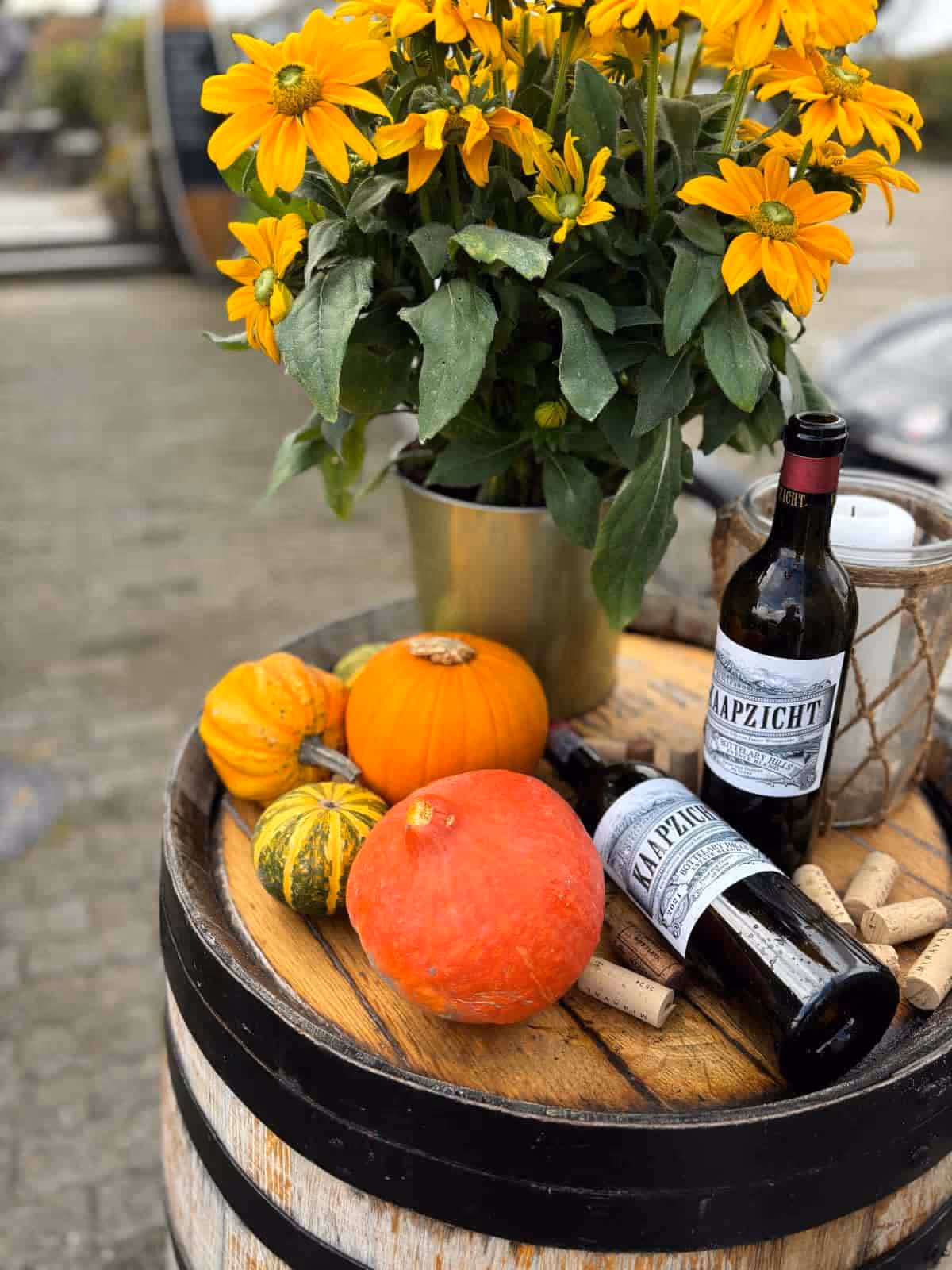 Herbstliche Dekoration mit Kürbissen, Sonnenblumen in Vase, zwei Weinflaschen und Weinkorken auf Fass.