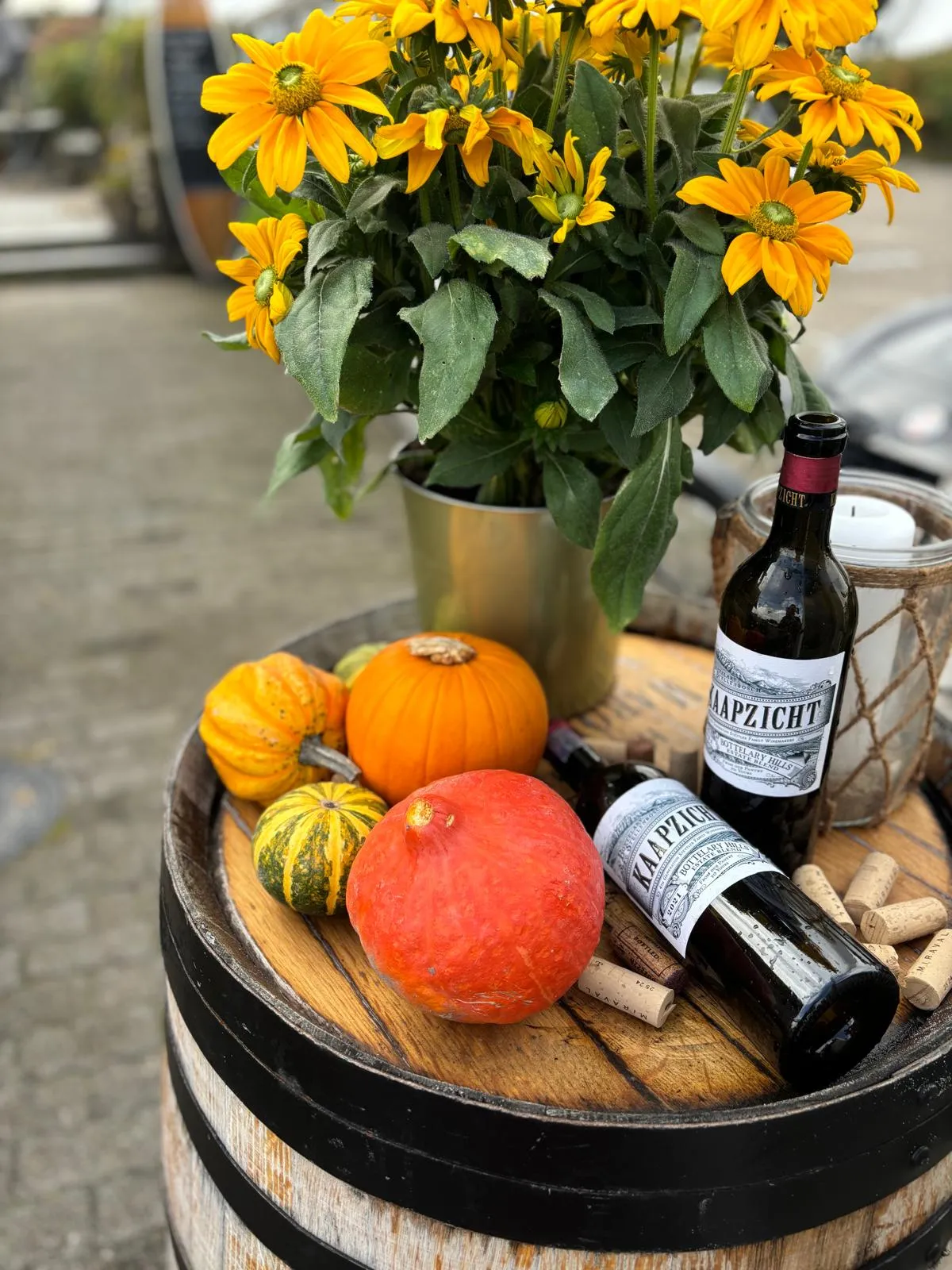 Herbstliche Dekoration mit Kürbissen, Sonnenblumen in Vase, zwei Weinflaschen und Weinkorken auf Fass.