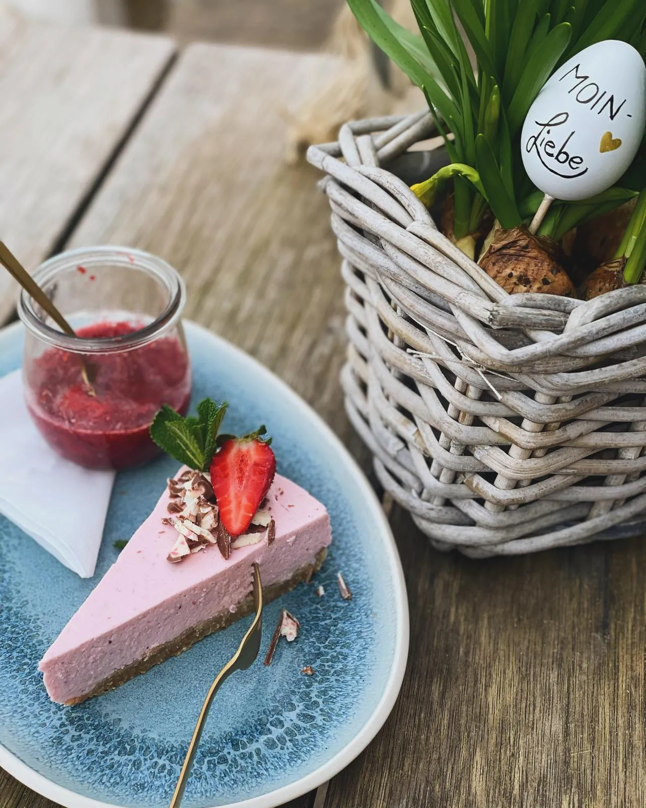 Ein Stück Erdbeerkuchen mit Erdbeersoße auf blauem Teller neben einem Korb mit grünen Pflanzen.