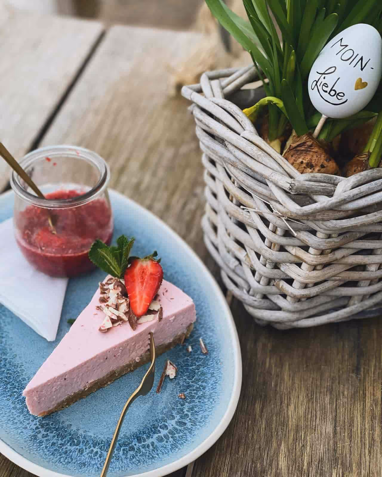 Ein Stück Erdbeerkuchen mit Erdbeersoße auf blauem Teller neben einem Korb mit grünen Pflanzen.