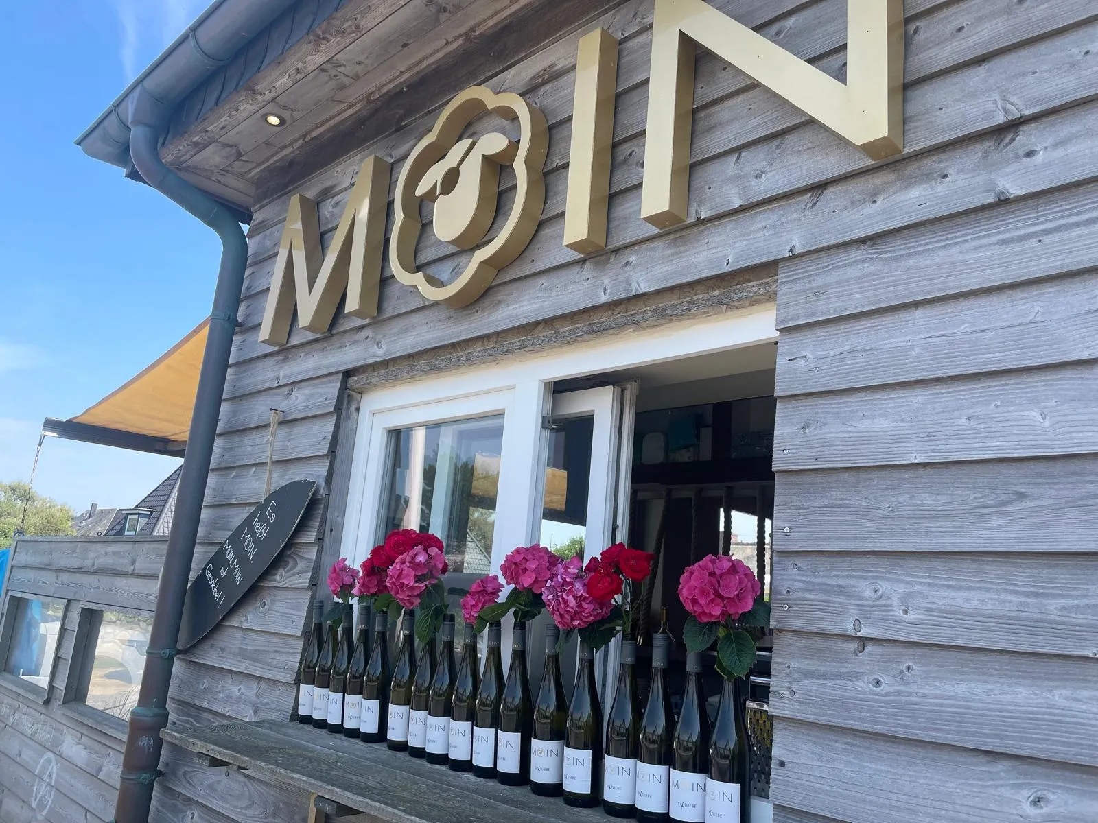 Reihen von Weinflaschen mit bunten Blumen vor einem Holzfenster unter einem goldenen MOIN-Schild.