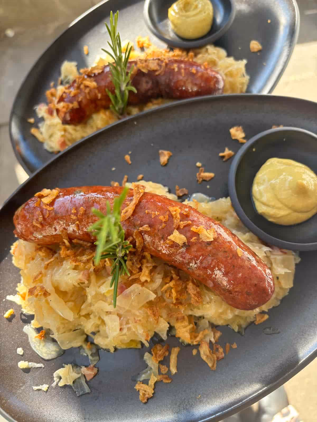 Gebratene Bratwurst auf Sauerkraut mit Röstzwiebeln, Rosmarin und Senf auf schwarzem Teller.