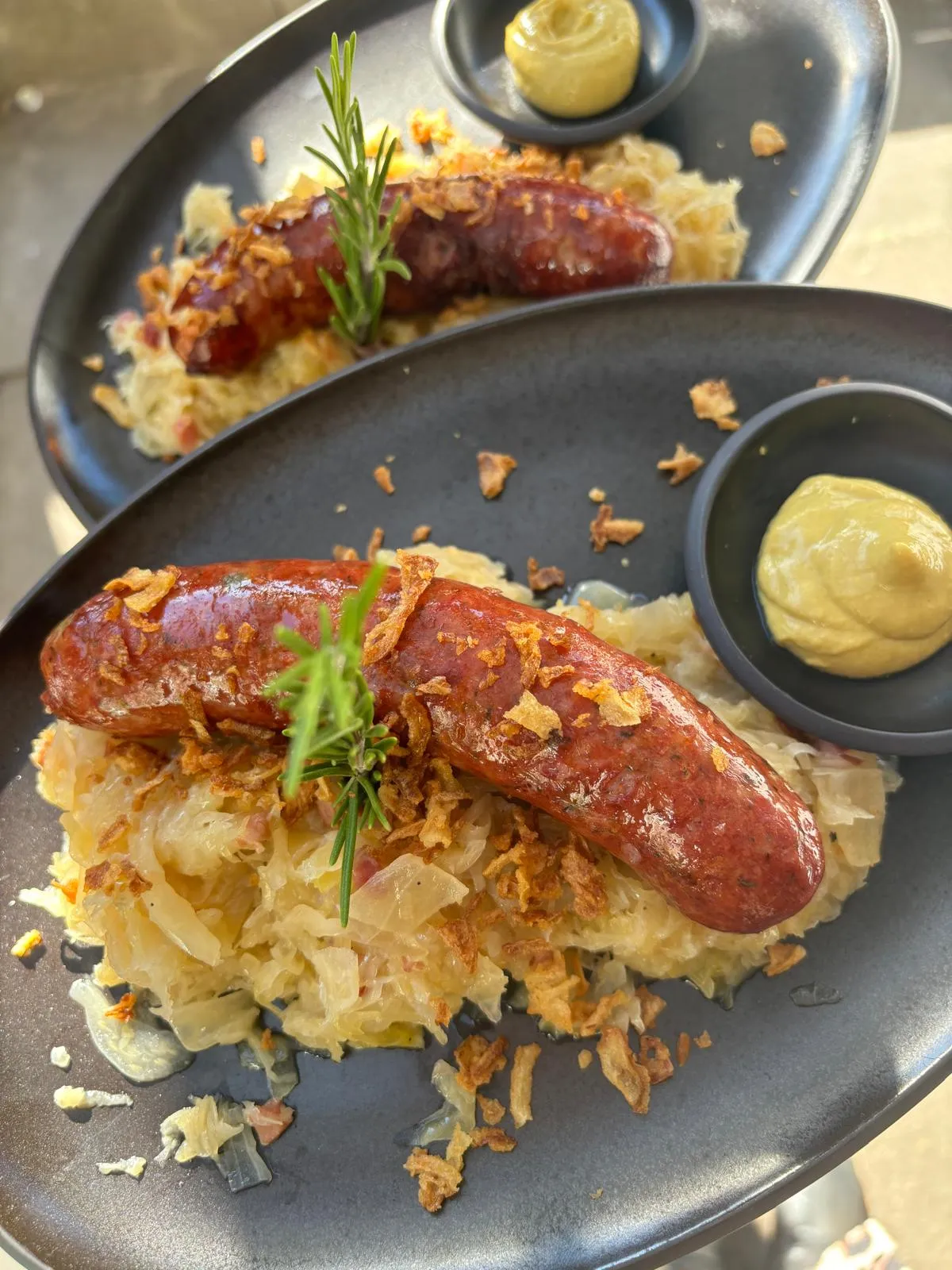 Gebratene Bratwurst auf Sauerkraut mit Röstzwiebeln, Rosmarin und Senf auf schwarzem Teller.