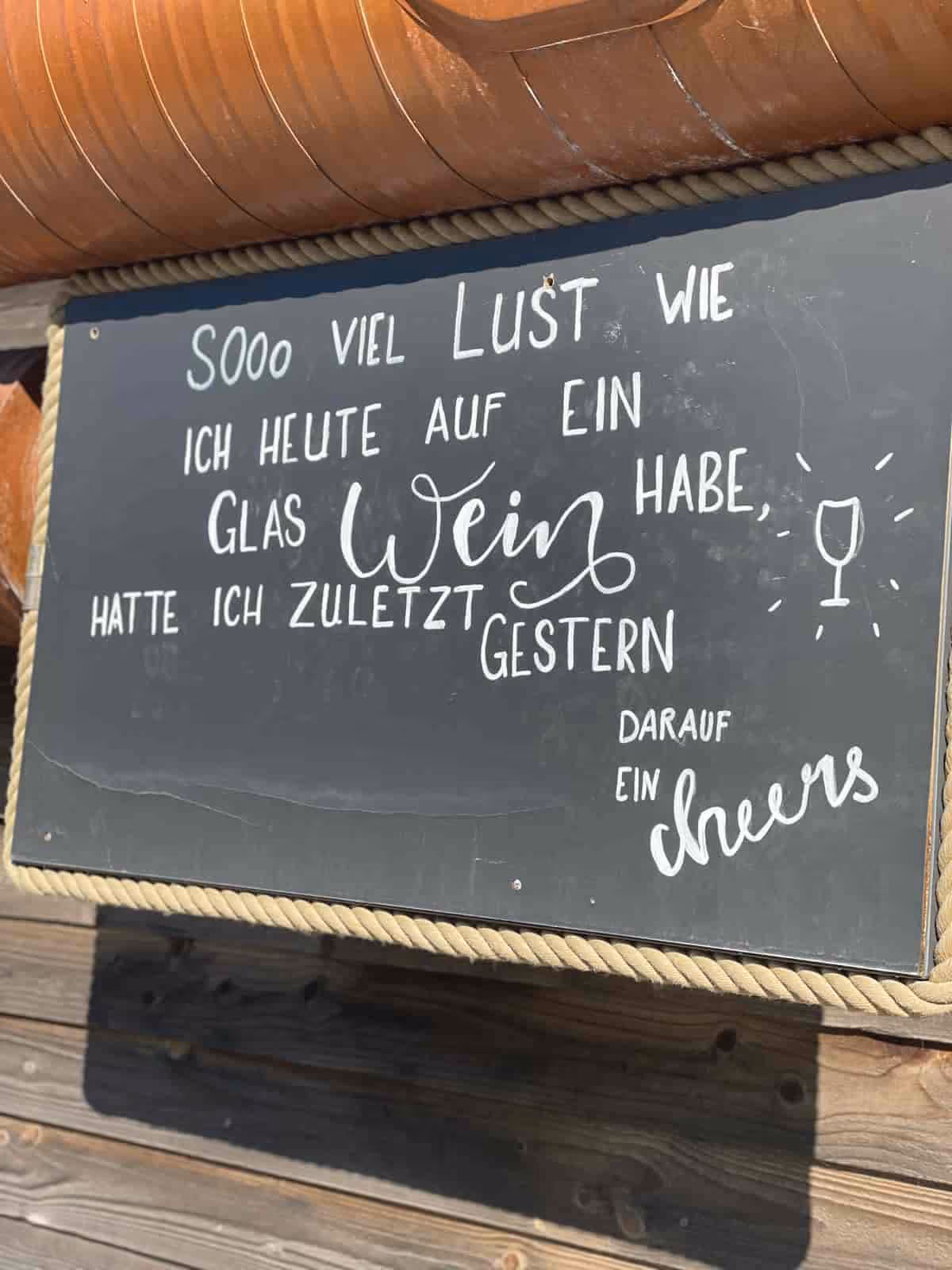 Schief hängende Tafel mit handgeschriebenem deutschen Spruch über Wein und Lust darauf.