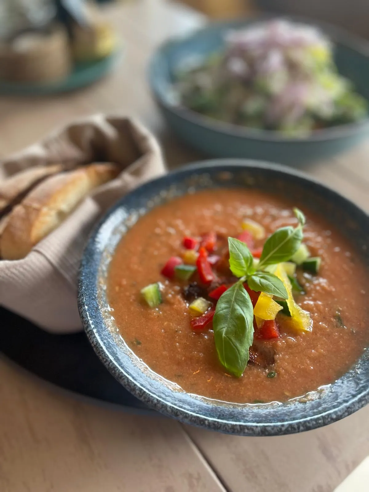 Schüssel mit Tomatensuppe garniert mit buntem Gemüse und Basilikum, dazu Brot im Hintergrund.