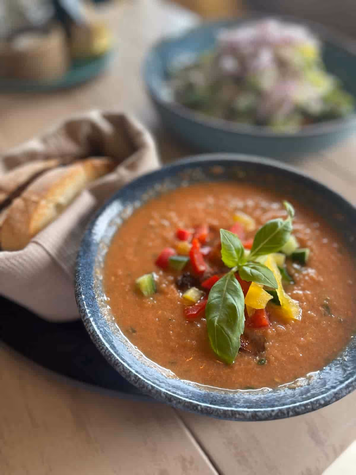 Schüssel mit Tomatensuppe garniert mit buntem Gemüse und Basilikum, dazu Brot im Hintergrund.