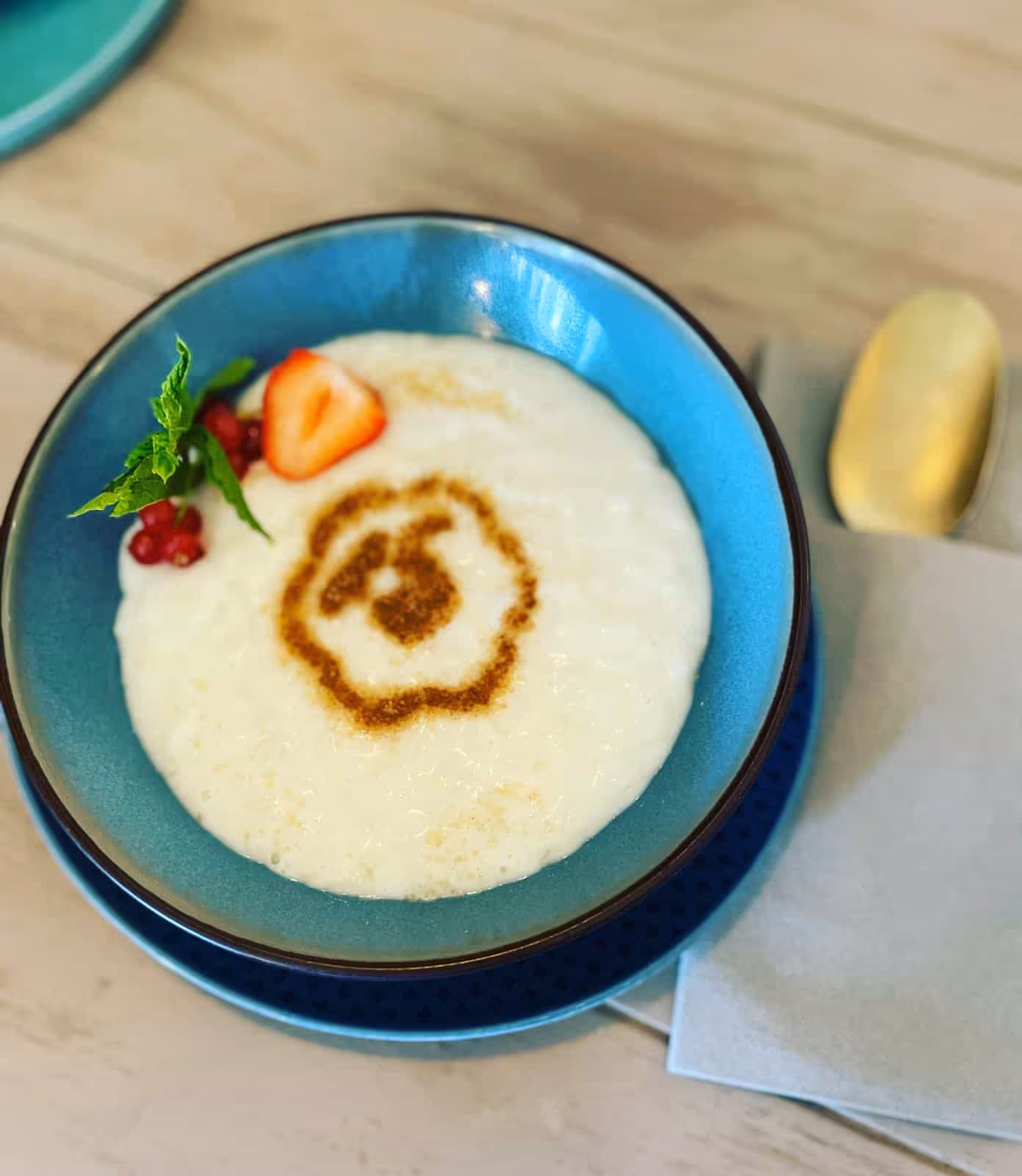 Blaue Schale mit Milchreis, Zimt, Erdbeere, roten Beeren und Minzblatt daneben Goldlöffel auf Serviette.
