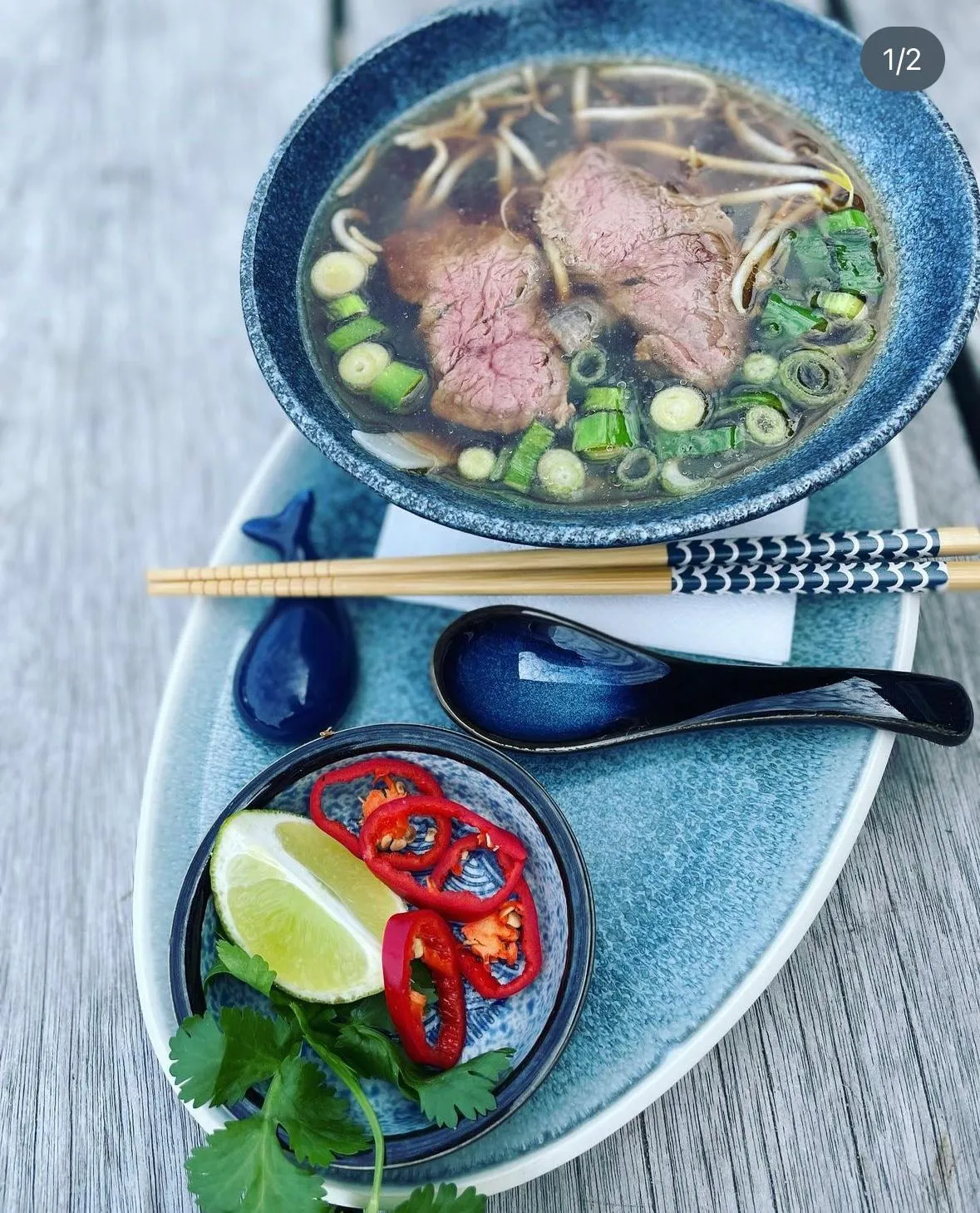 Schale mit Pho-Suppe, Rindfleisch, Frühlingszwiebeln, Limette, Chili und Koriander, Essstäbchen und Löffel.