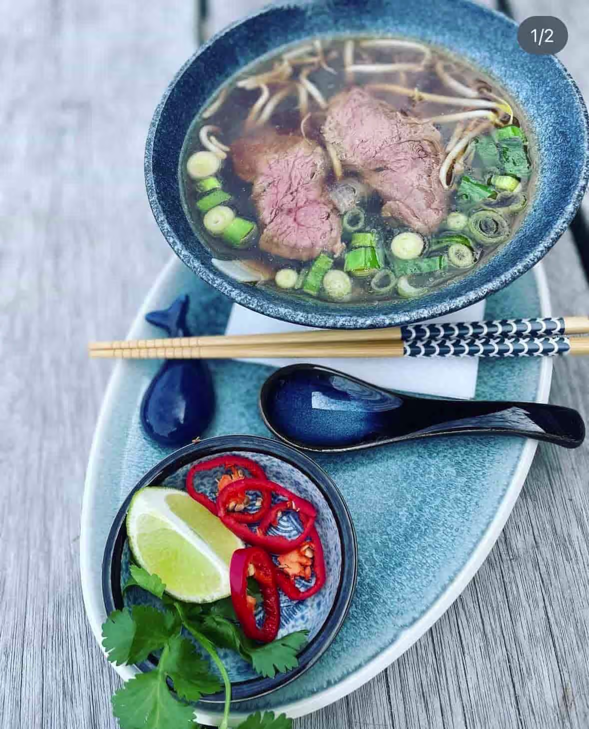 Schale mit Pho-Suppe, Rindfleisch, Frühlingszwiebeln, Limette, Chili und Koriander, Essstäbchen und Löffel.