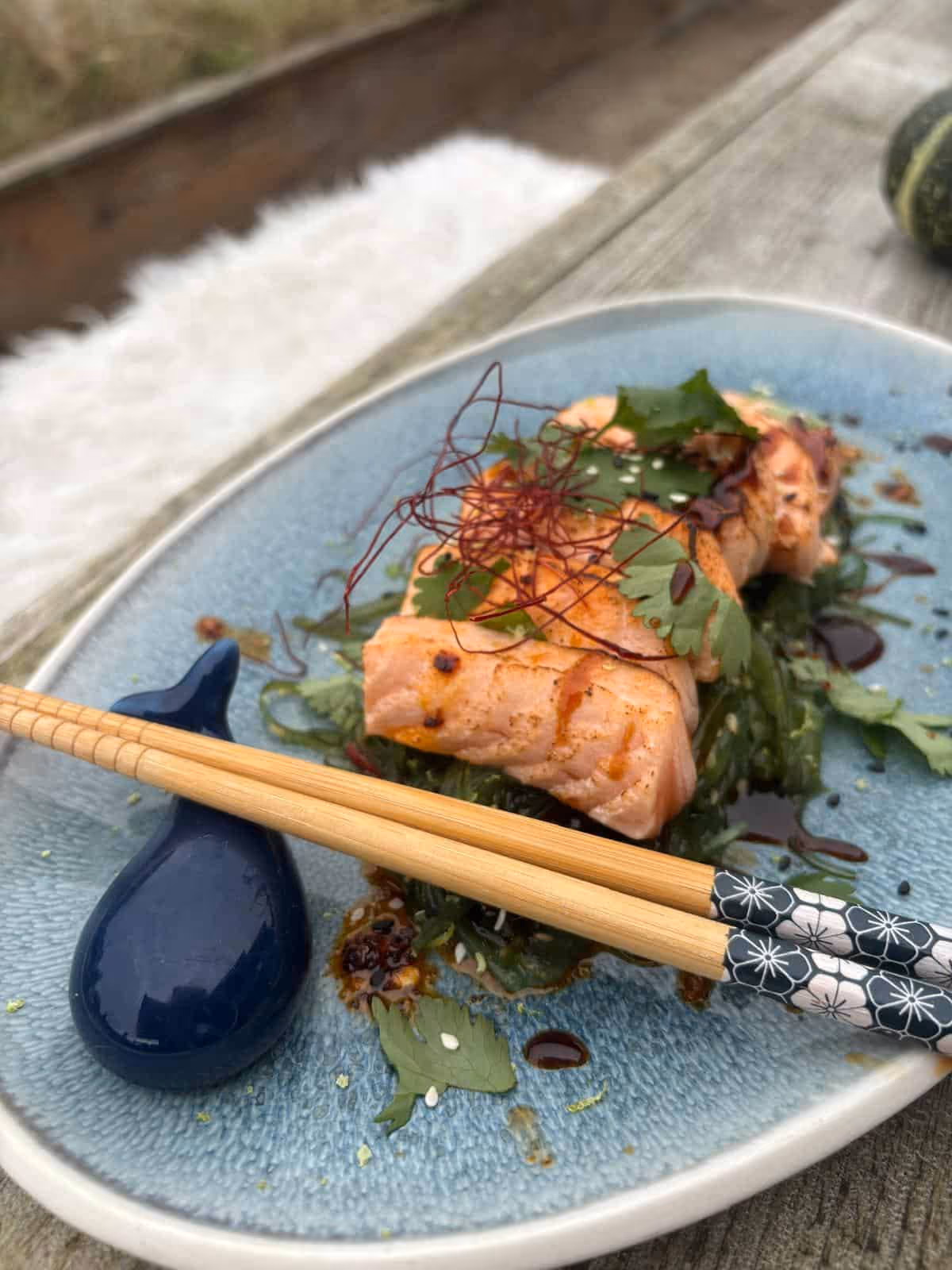Gegrilltes Lachsstück auf Salat mit Sojasauce, Koriander und Stäbchen auf blauem Teller.