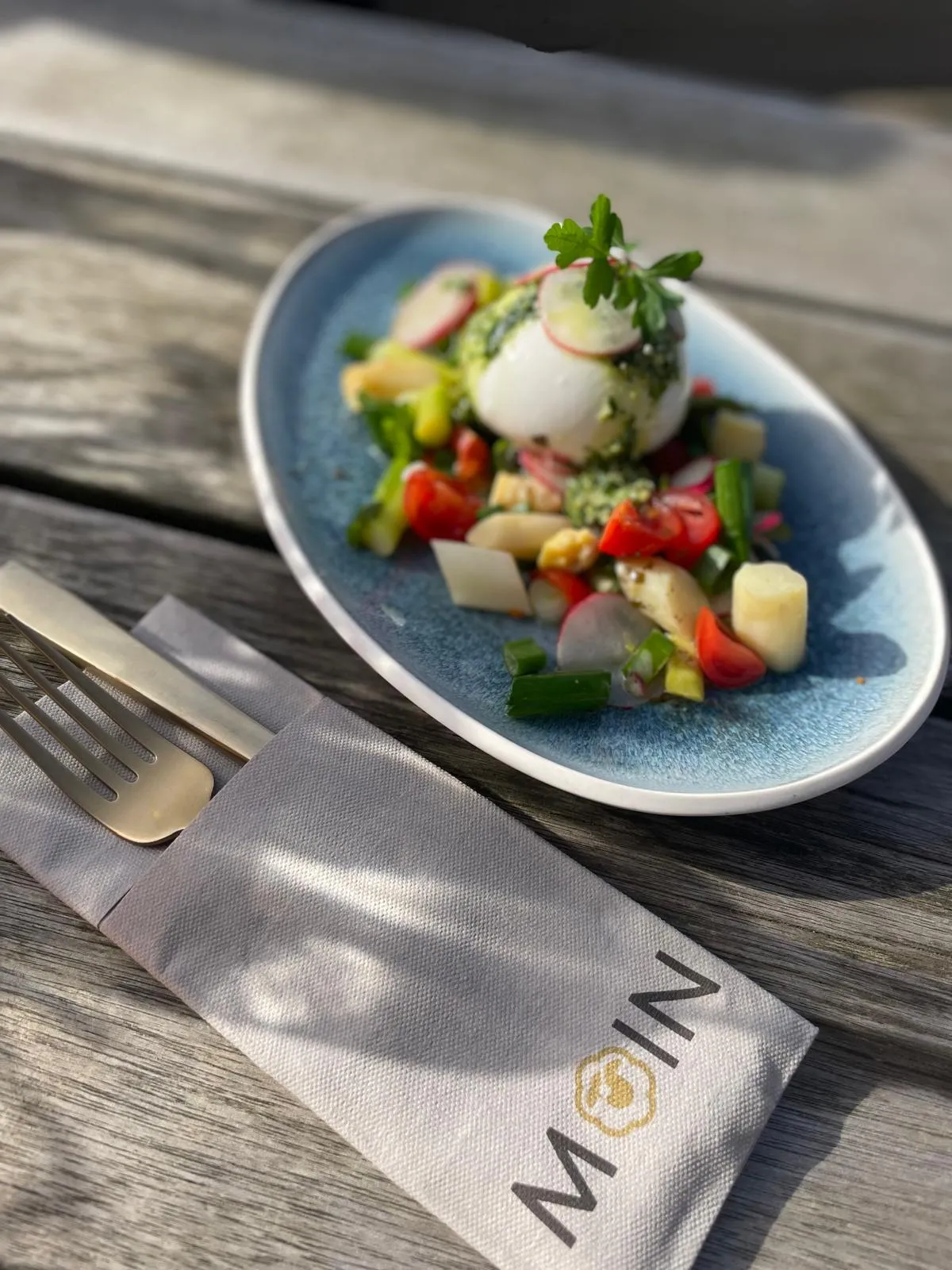 Burrata mit frischem Gemüse und Kräutern auf blauem Teller neben Besteck auf Holzbank.