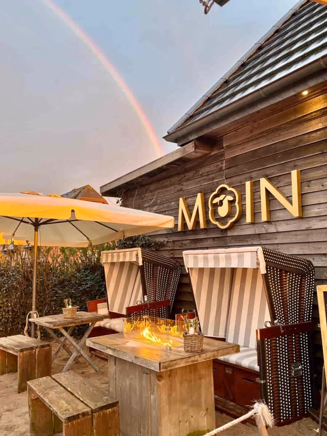 Gemütliche Sitzplätze mit Strandkörben und Feuerstelle vor Holzhaus mit MOIN-Schrift und Regenbogen.