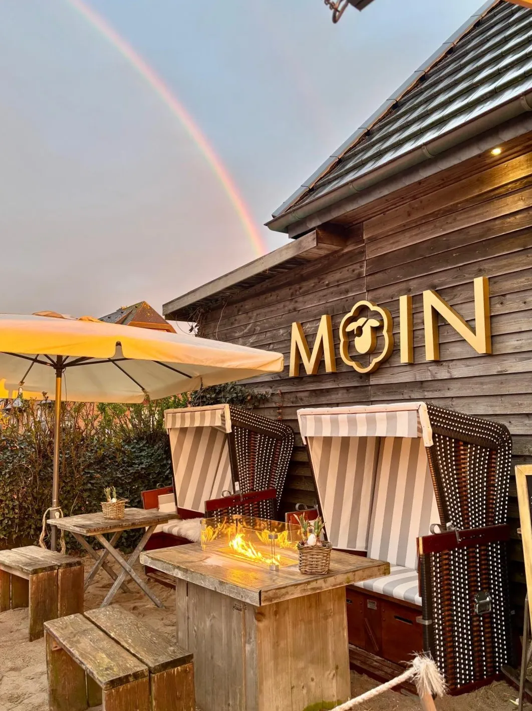 Gemütliche Sitzplätze mit Strandkörben und Feuerstelle vor Holzhaus mit MOIN-Schrift und Regenbogen.