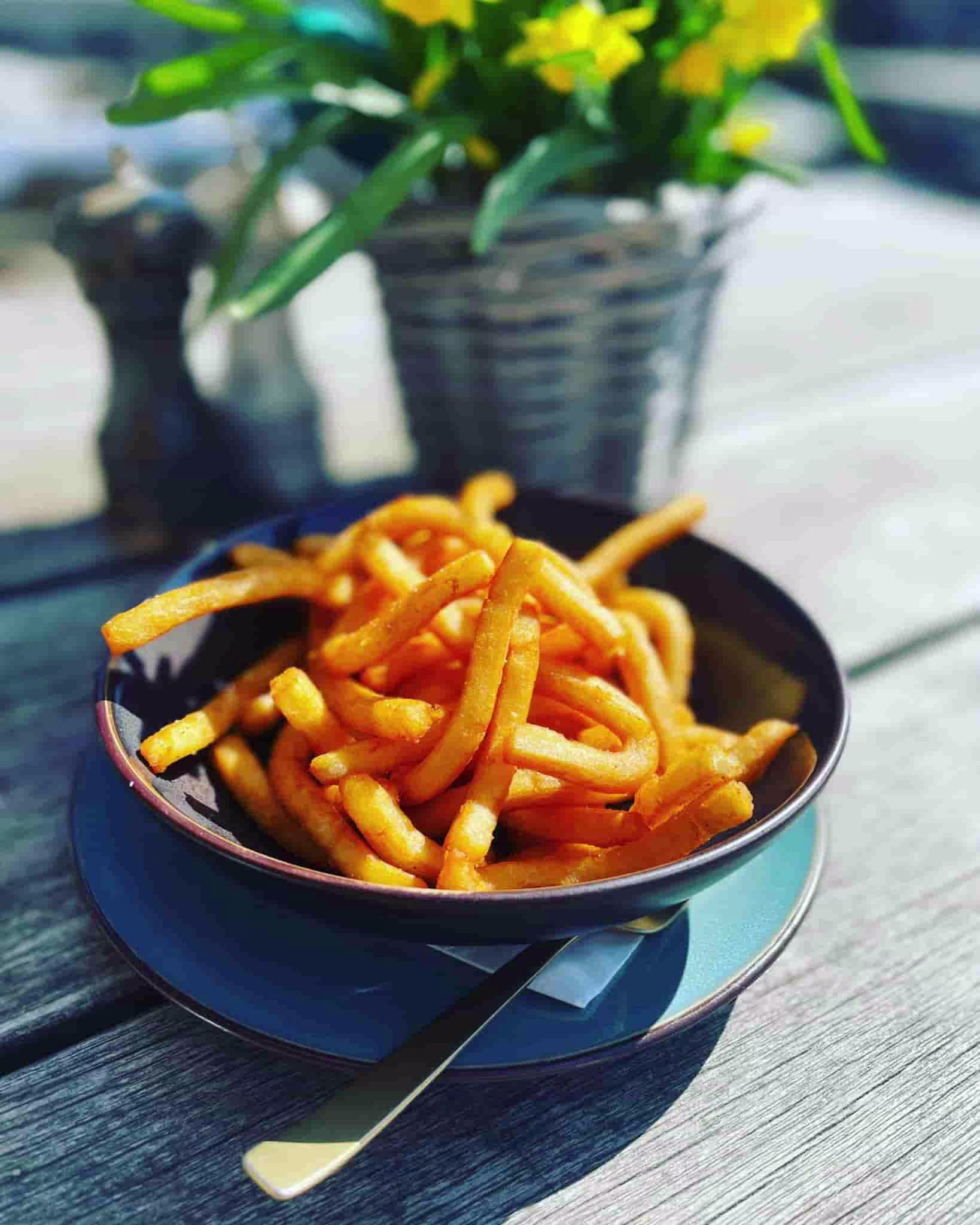 Schwarze Schüssel mit knusprigen, goldenen Pommes auf blauem Teller mit Messer auf Holzterrasse.