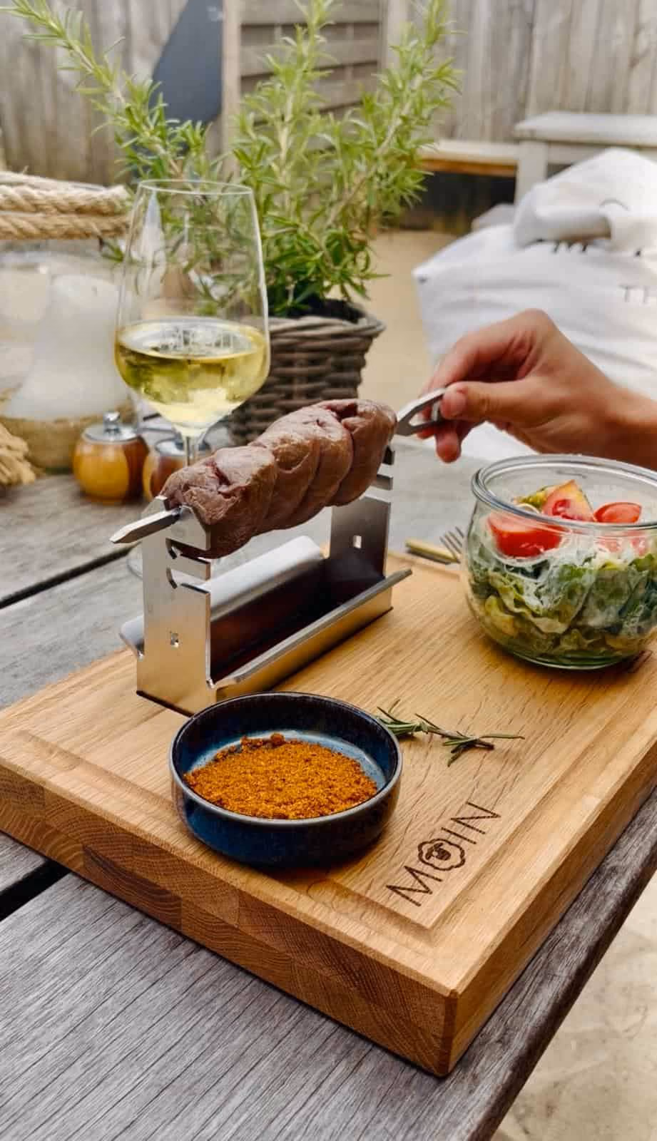Hand hält Grillgabel mit Fleischstück über kleinem Edelstahl-Tischgrill neben Salat und Gewürz.