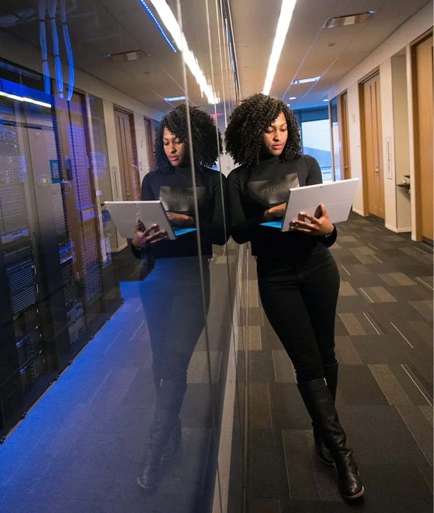Woman with curly hair in black outfit leaning against a glass wall in a server room hallway, working on a laptop.