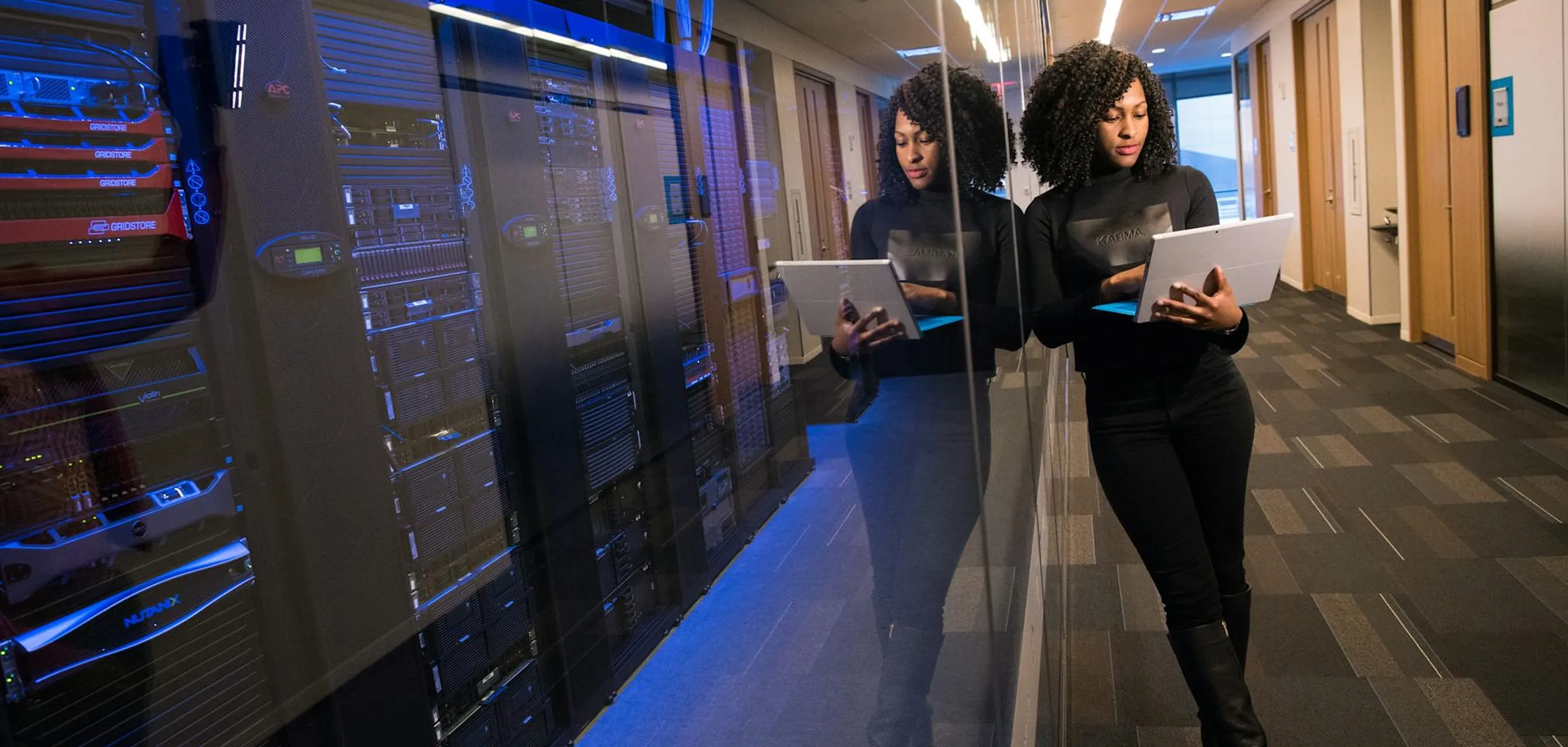 Woman in black outfit using a laptop while standing in a hallway beside server racks with blue lighting.