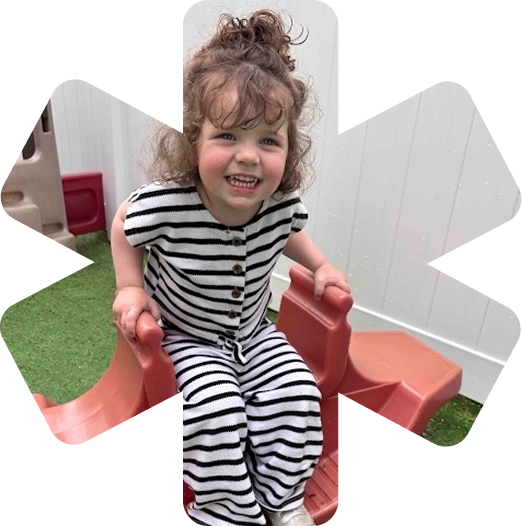 Smiling toddler with curly hair in a black and white striped outfit sitting on a red plastic slide outdoors.