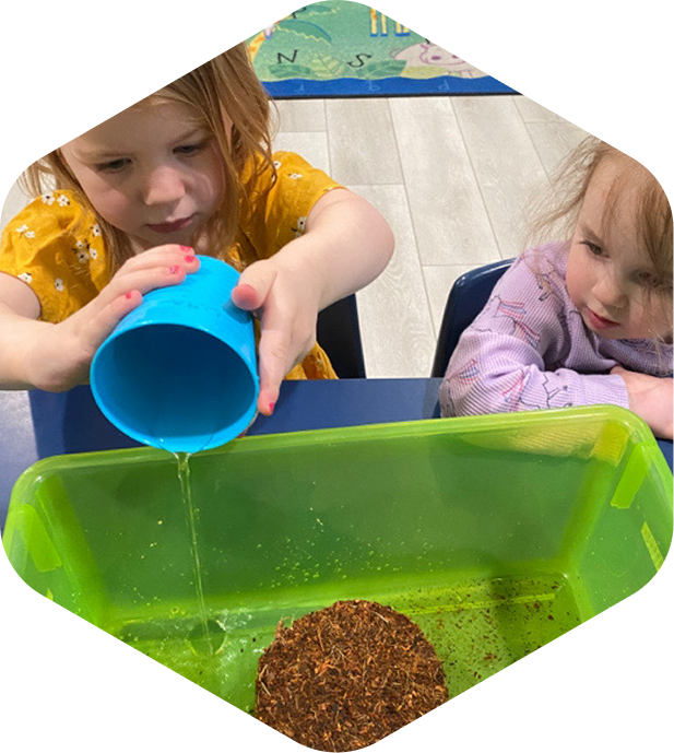 Child in yellow dress pouring water from a blue cup into a green container with soil, while another child watches.
