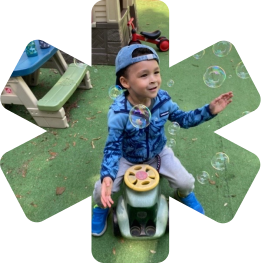 Young boy in a blue jacket and cap sitting on a toy car outdoors, reaching for floating soap bubbles.