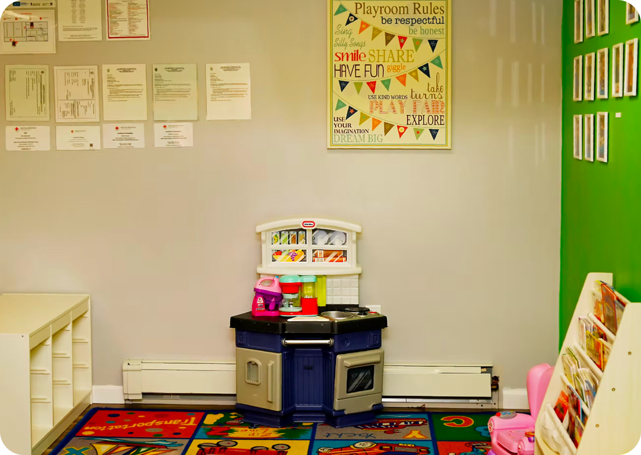 Bright daycare room with colorful animal-themed rugs, wooden cribs, shelves of toys, and a play kitchen set.