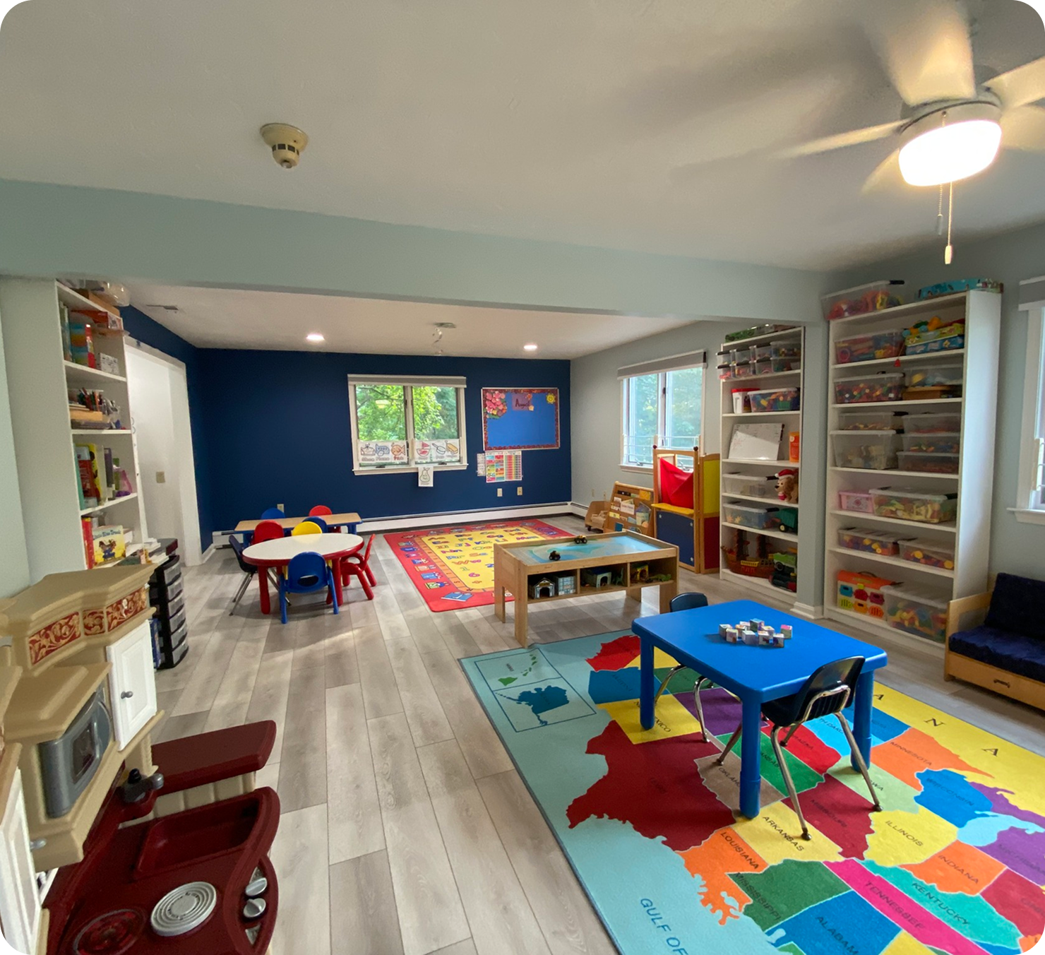 Bright classroom with colorful small tables and chairs, educational rugs, shelves filled with toys and learning materials, and large windows.
