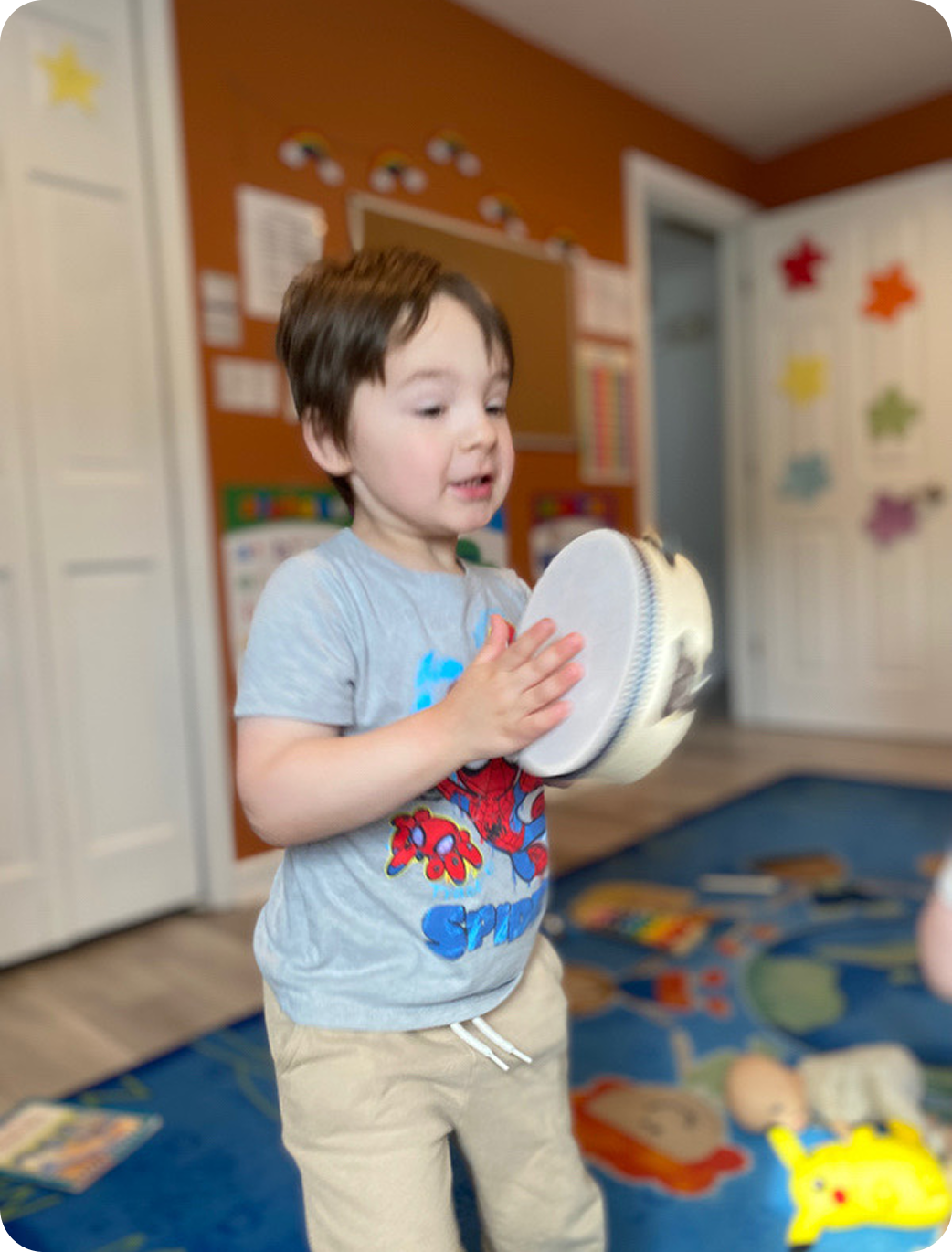 Young child wearing a Spiderman t-shirt playing a tambourine in a colorful room with toys and educational posters.