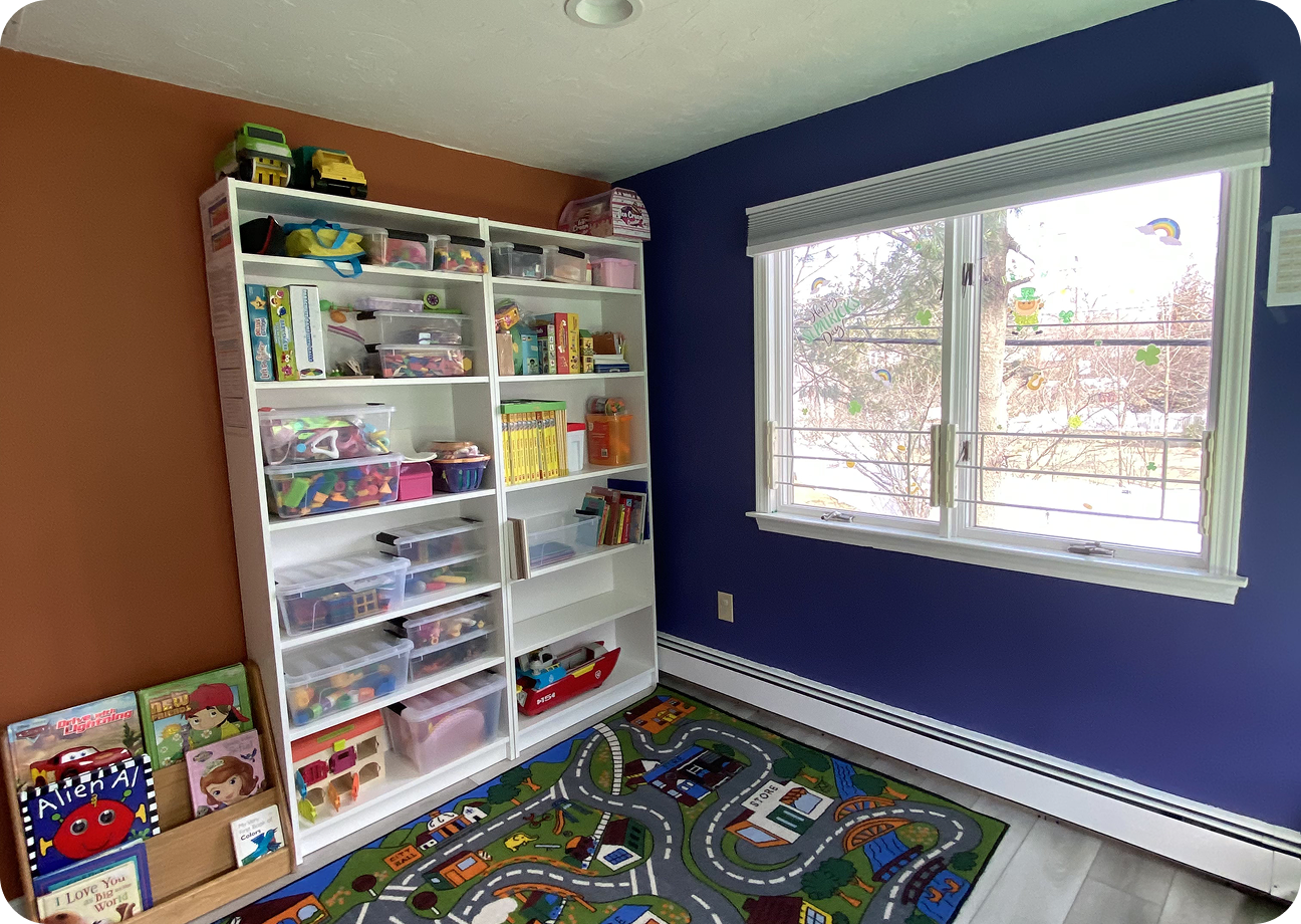 Bright and organized daycare room with cribs, rocking chair, colorful play mats, toys, and cabinets under warm lighting.