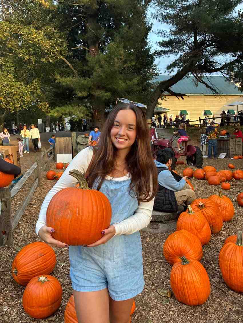 Smiling young woman with long brown hair holding a large orange pumpkin, wearing a white shirt and blue overalls.