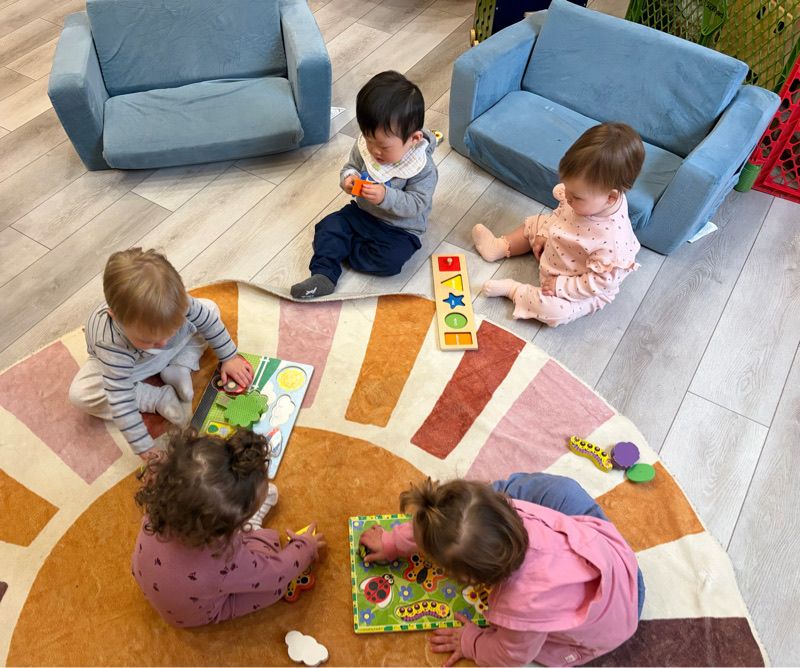 Bright daycare room with colorful animal-themed rugs, wooden cribs, shelves of toys, and a play kitchen set.
