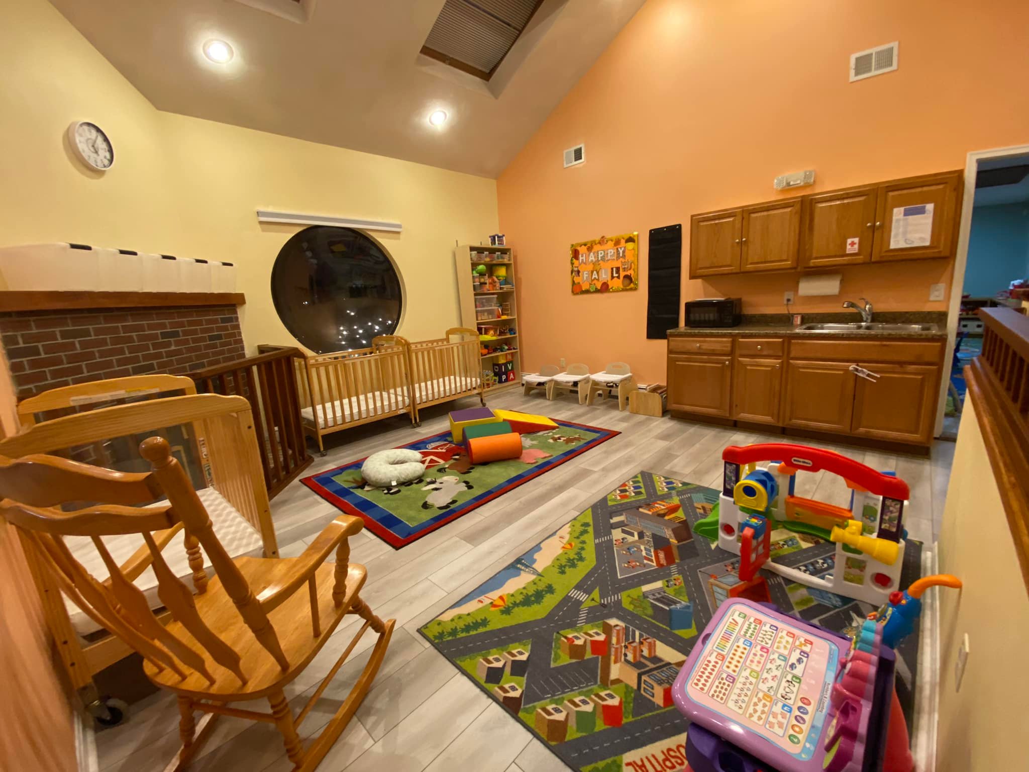 Bright daycare room with colorful animal-themed rugs, wooden cribs, shelves of toys, and a play kitchen set.