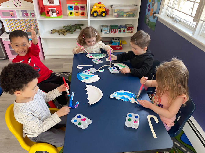 Bright daycare room with colorful animal-themed rugs, wooden cribs, shelves of toys, and a play kitchen set.