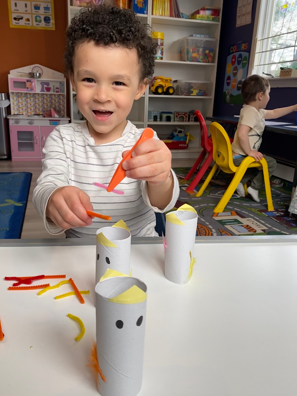 Bright daycare room with wooden cribs, colorful animal-themed rugs, a baby play gym, toy kitchen, and shelves filled with toys.