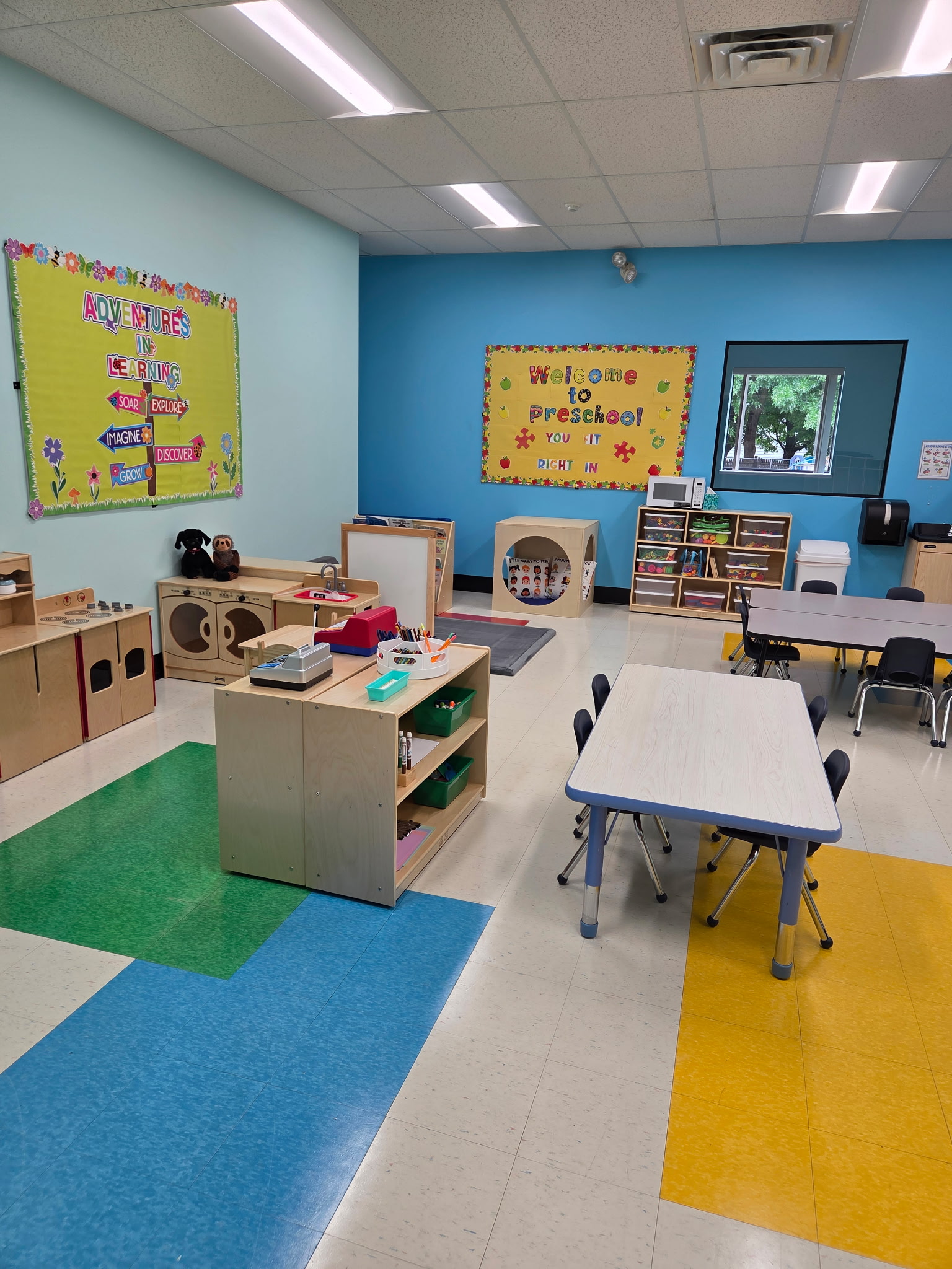 Bright and organized daycare room with cribs, rocking chair, colorful play mats, toys, and cabinets under warm lighting.