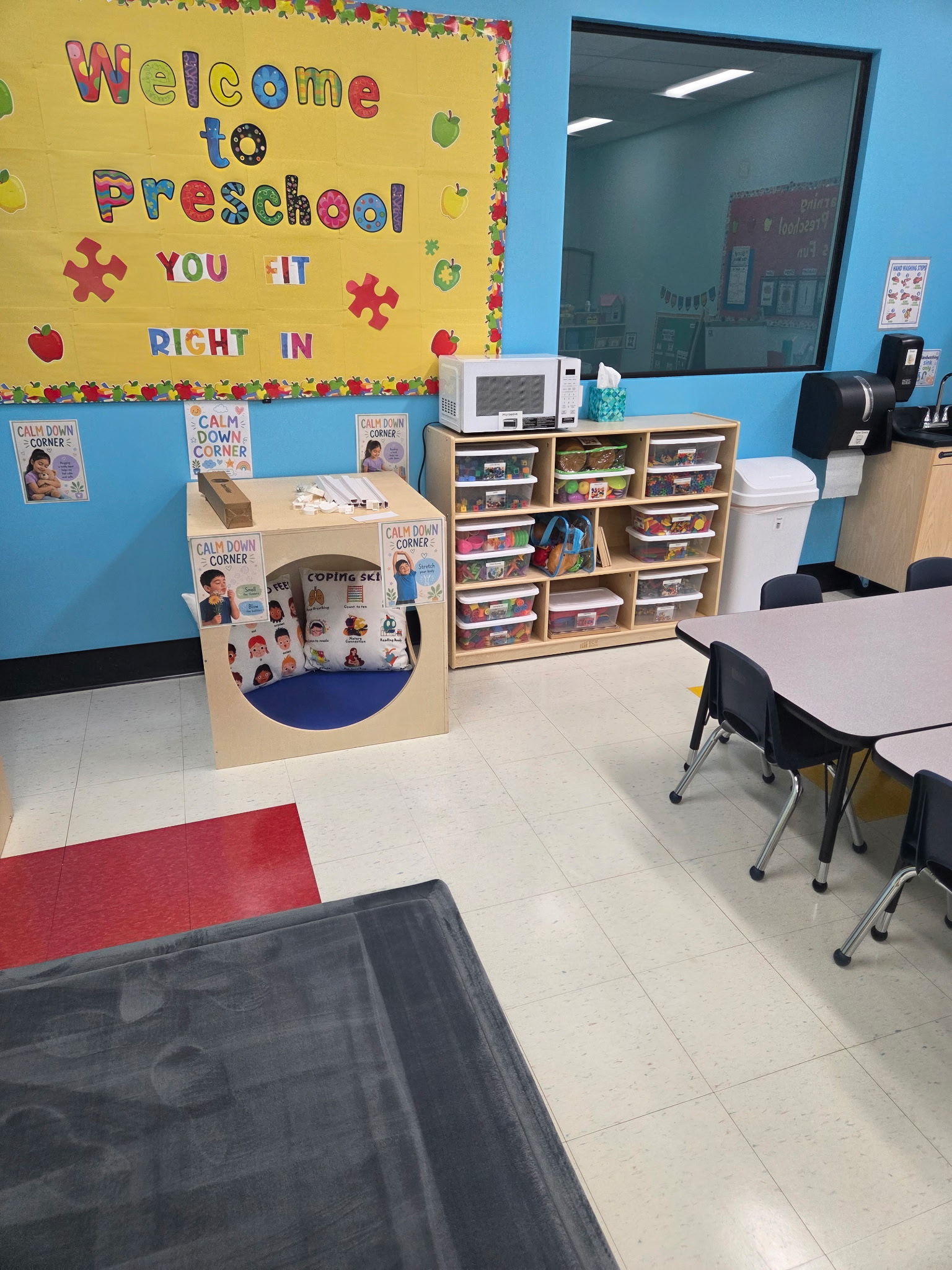 Bright daycare room with colorful animal-themed rugs, wooden cribs, shelves of toys, and a play kitchen set.
