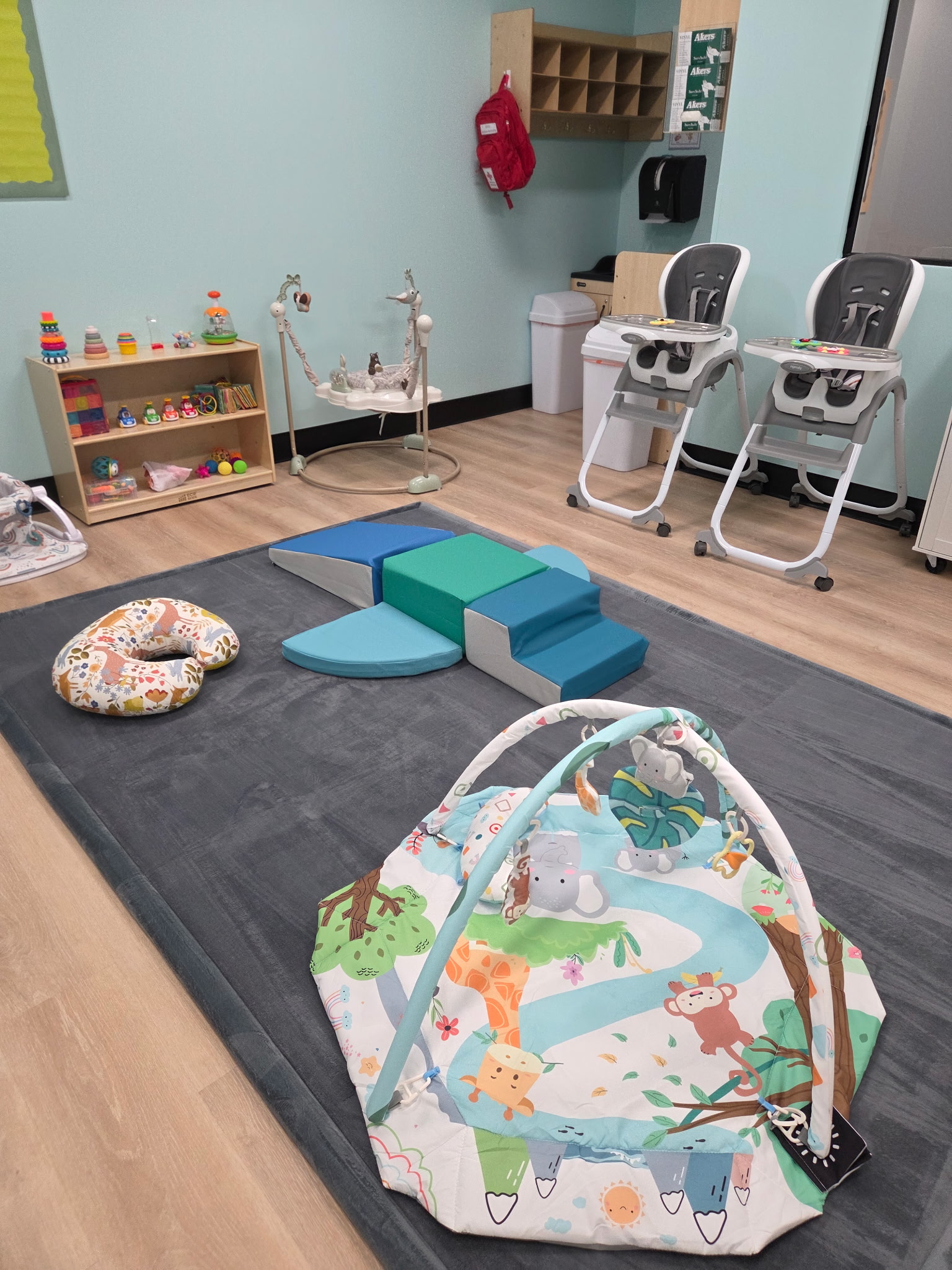 Children's playroom area with a toy kitchen set, blue baby seat, and a tall shelf filled with organized colorful toys and educational blocks next to foam climbing blocks.