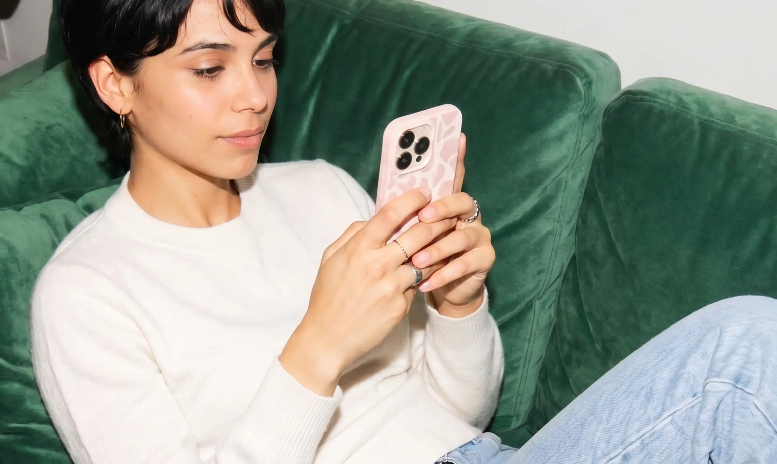 Young woman in a white sweater looking at her phone on a green velvet sofa