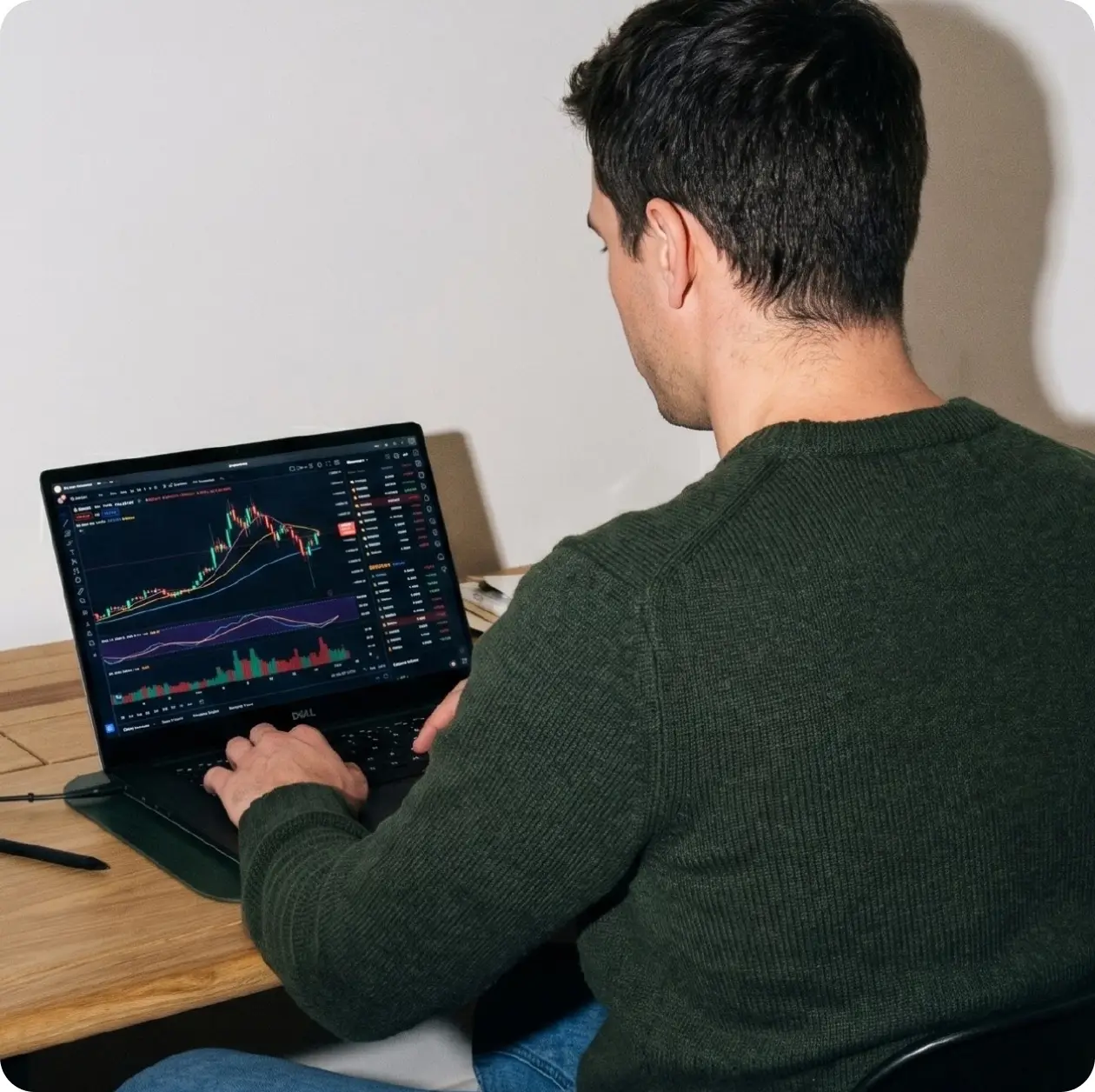 Man in a green sweater viewing crypto trading charts on a laptop at his desk