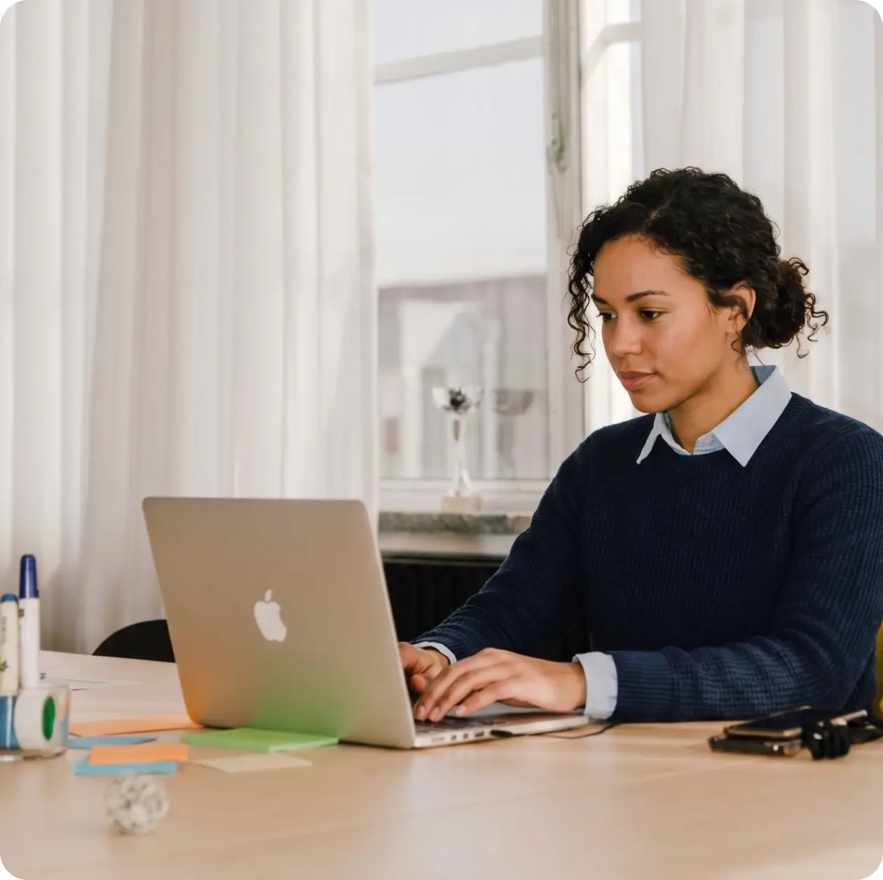 Young woman in a navy sweater focused on work at her laptop in a bright office