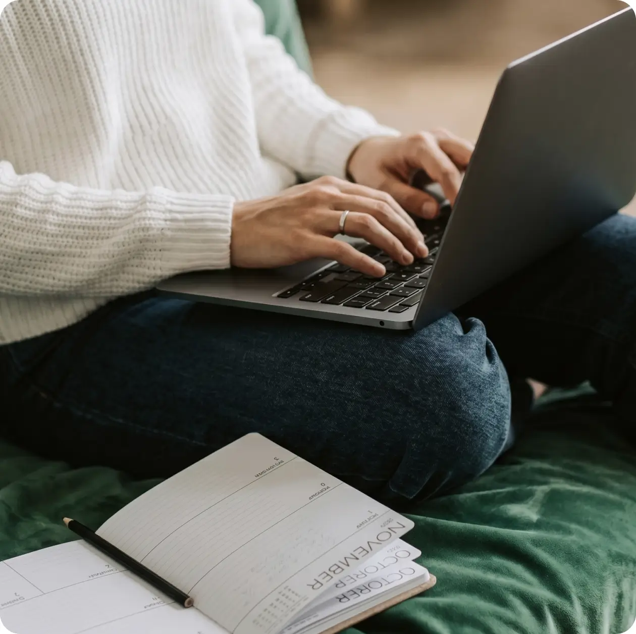 Person in a white sweater typing on a laptop with an open planner notebook nearby