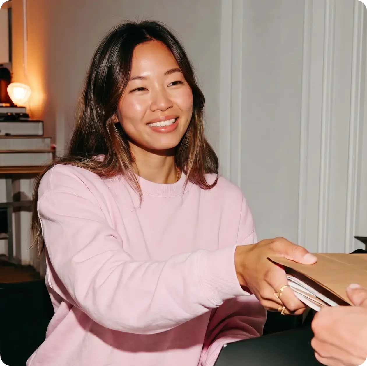 Smiling young woman in a pink sweatshirt handing over documents