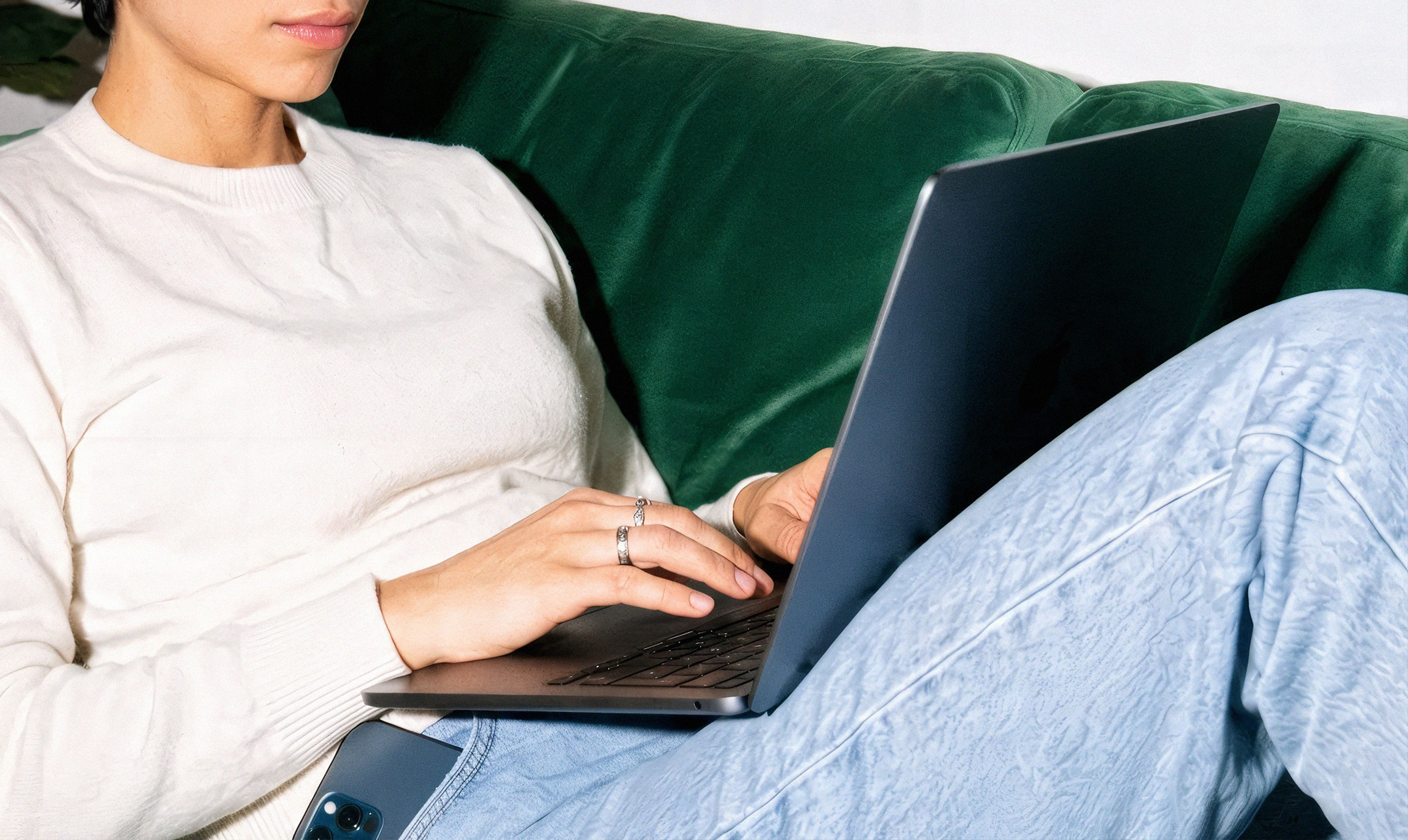 Young woman with short hair in a white sweater working on a laptop while relaxing on a green velvet sofa