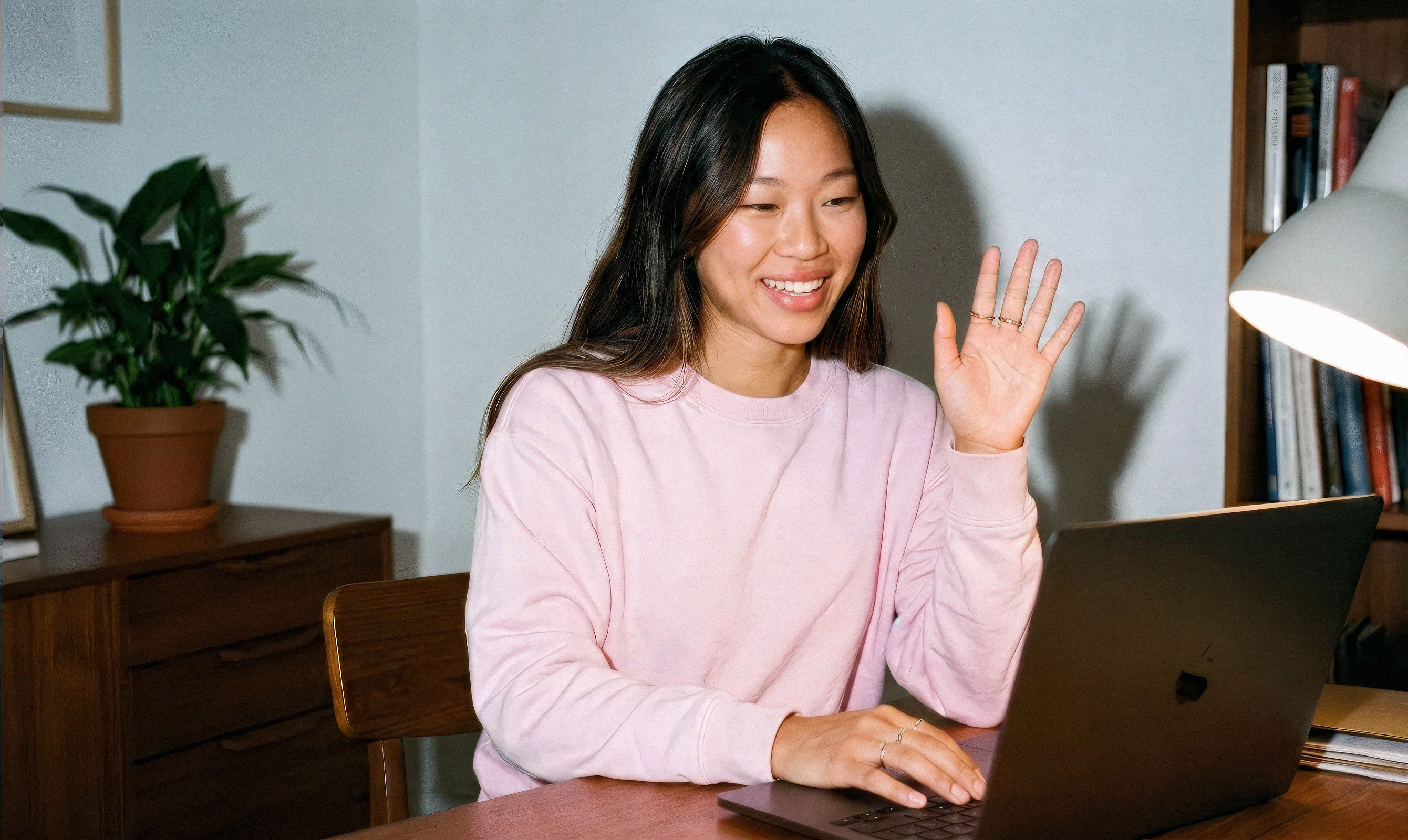 Smiling young woman in a pink sweatshirt waving during a video call on her laptop at a home desk