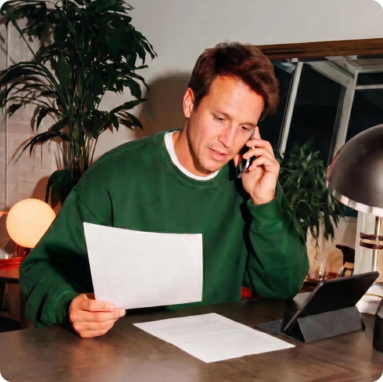 Man in a green sweatshirt reviewing a document while on a phone call at his desk
