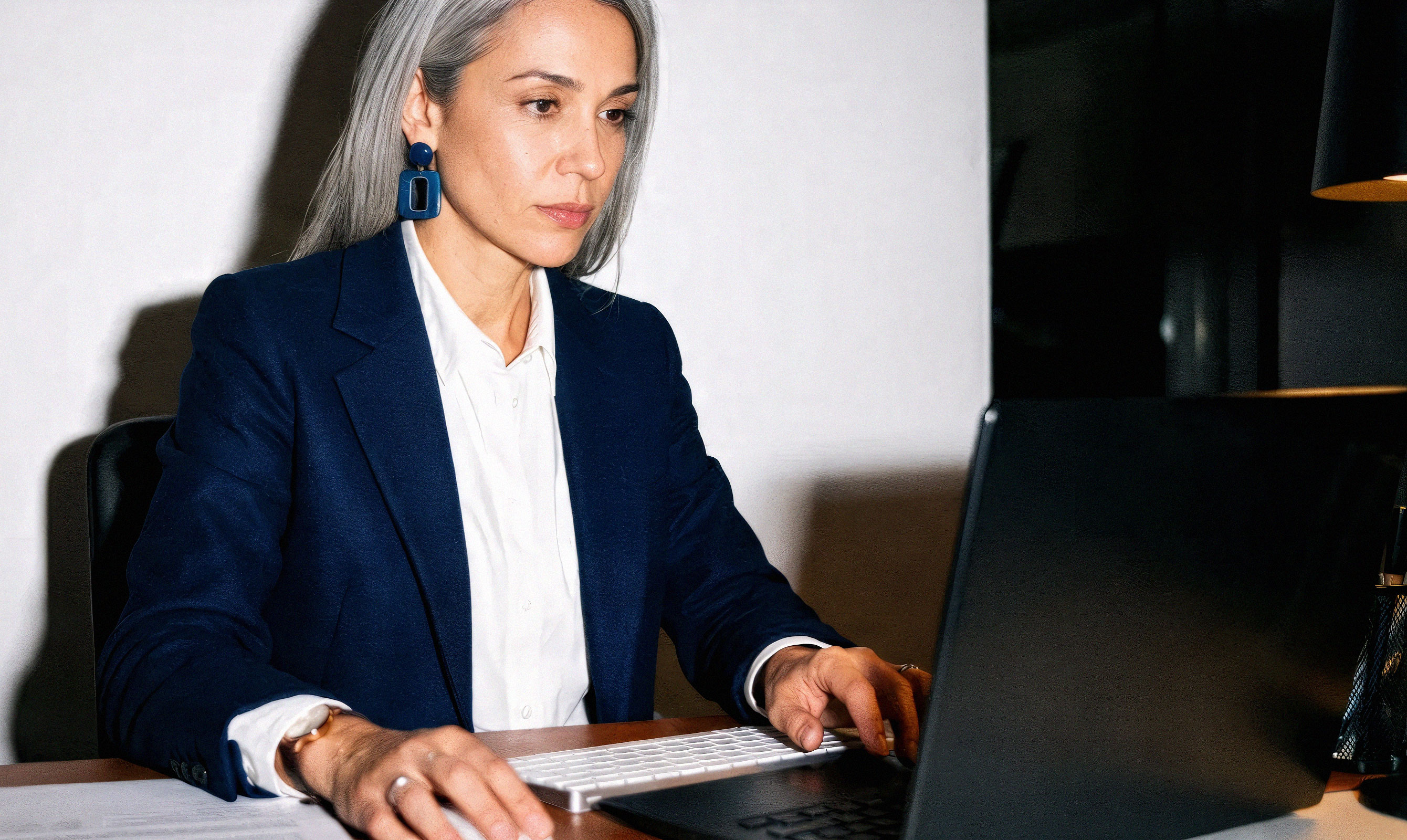 Professional woman with grey hair in a navy blazer focused on work at her laptop