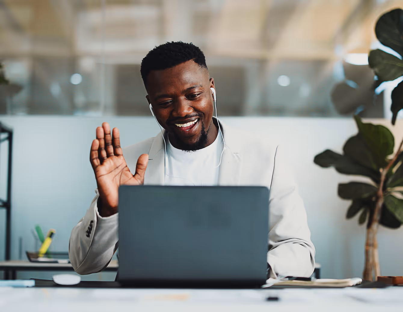 Smiling man wearing earphones waving at a laptop during a video call in a modern office.