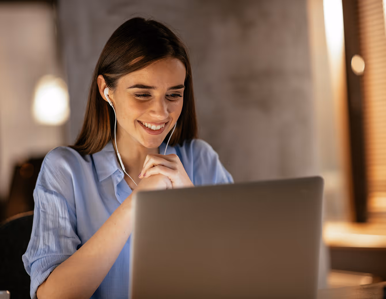 Smiling young woman wearing earphones, looking at a laptop screen in a cozy indoor setting.