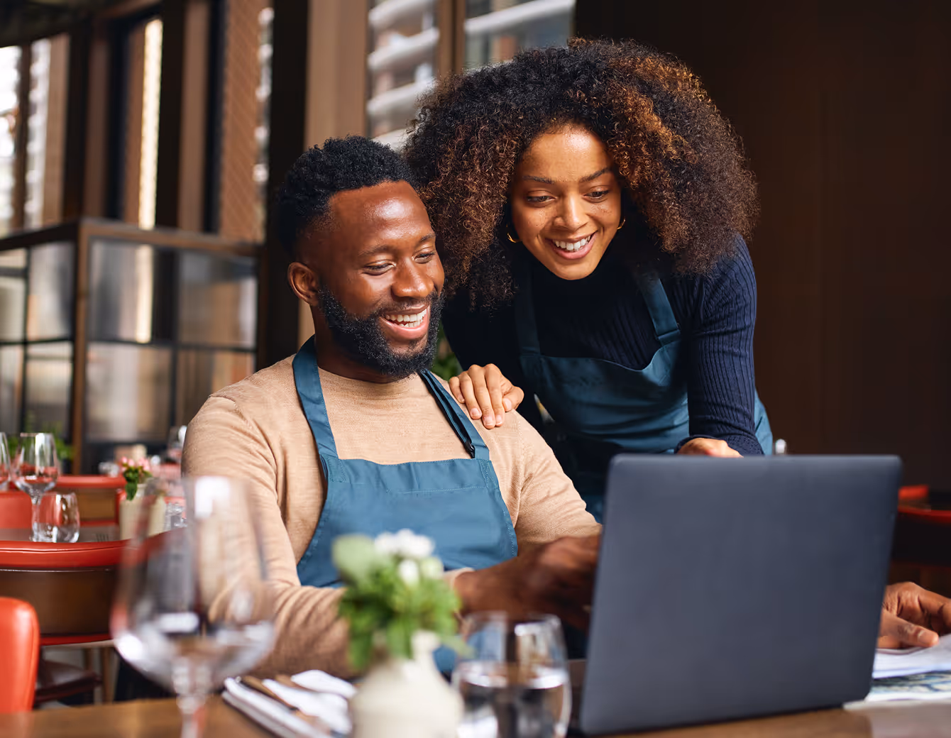 Smiling man and woman wearing aprons looking at a laptop in a restaurant setting.