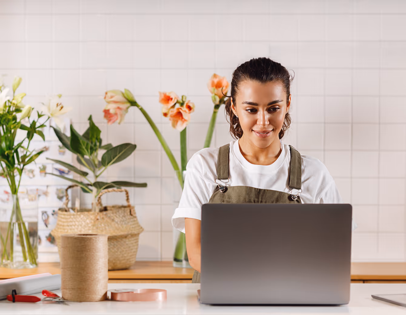 Smiling woman in a white shirt and green apron working on a laptop at a table with plants and crafting materials in the background.